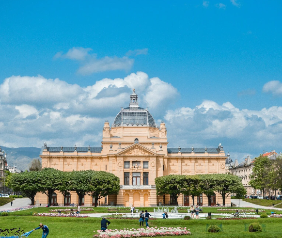 A large domed roof building in Croatia, surrounded by a green park and people walking.