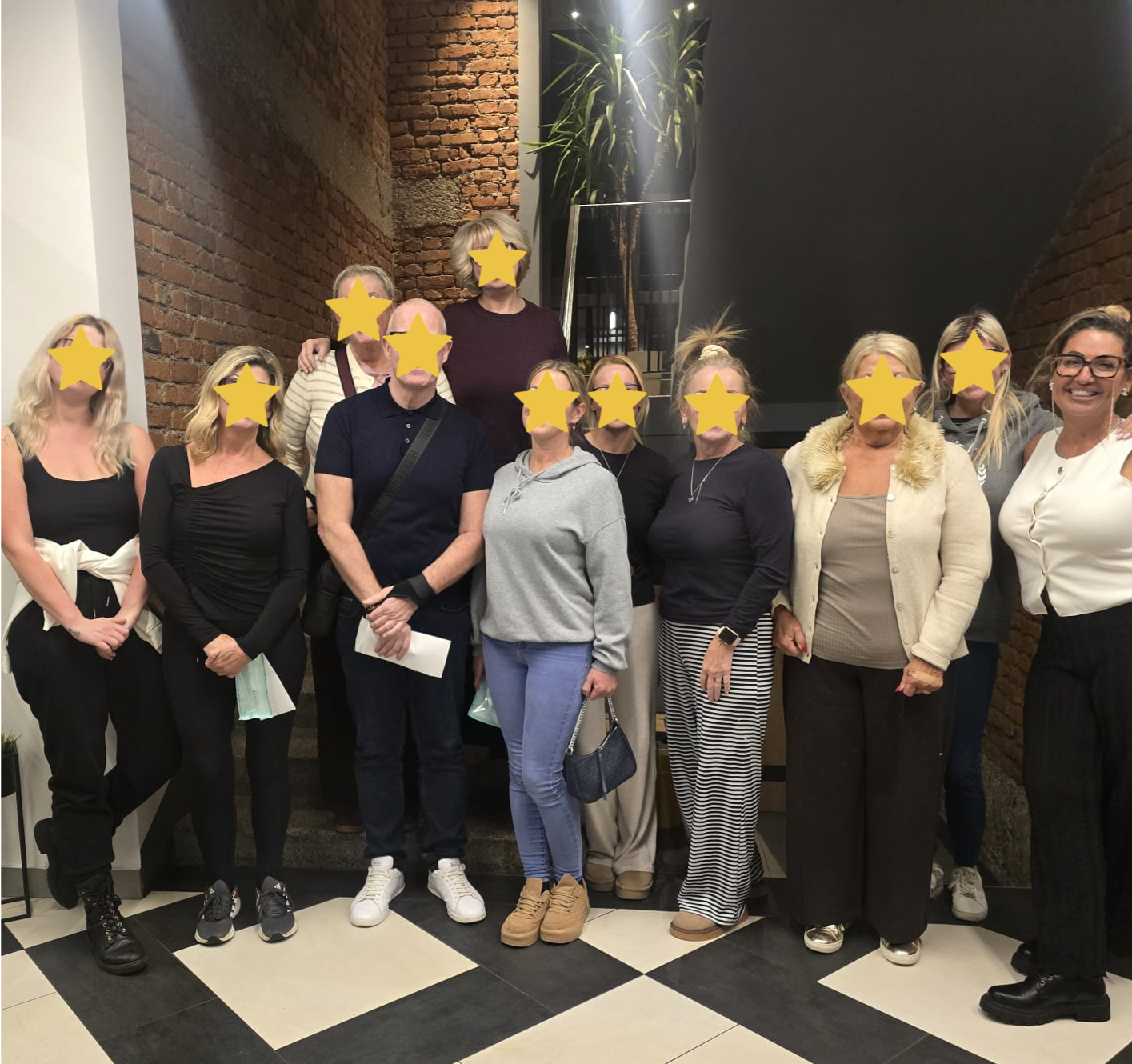 Group of twelve people standing together indoors, some smiling, in front of brick and dark walls.