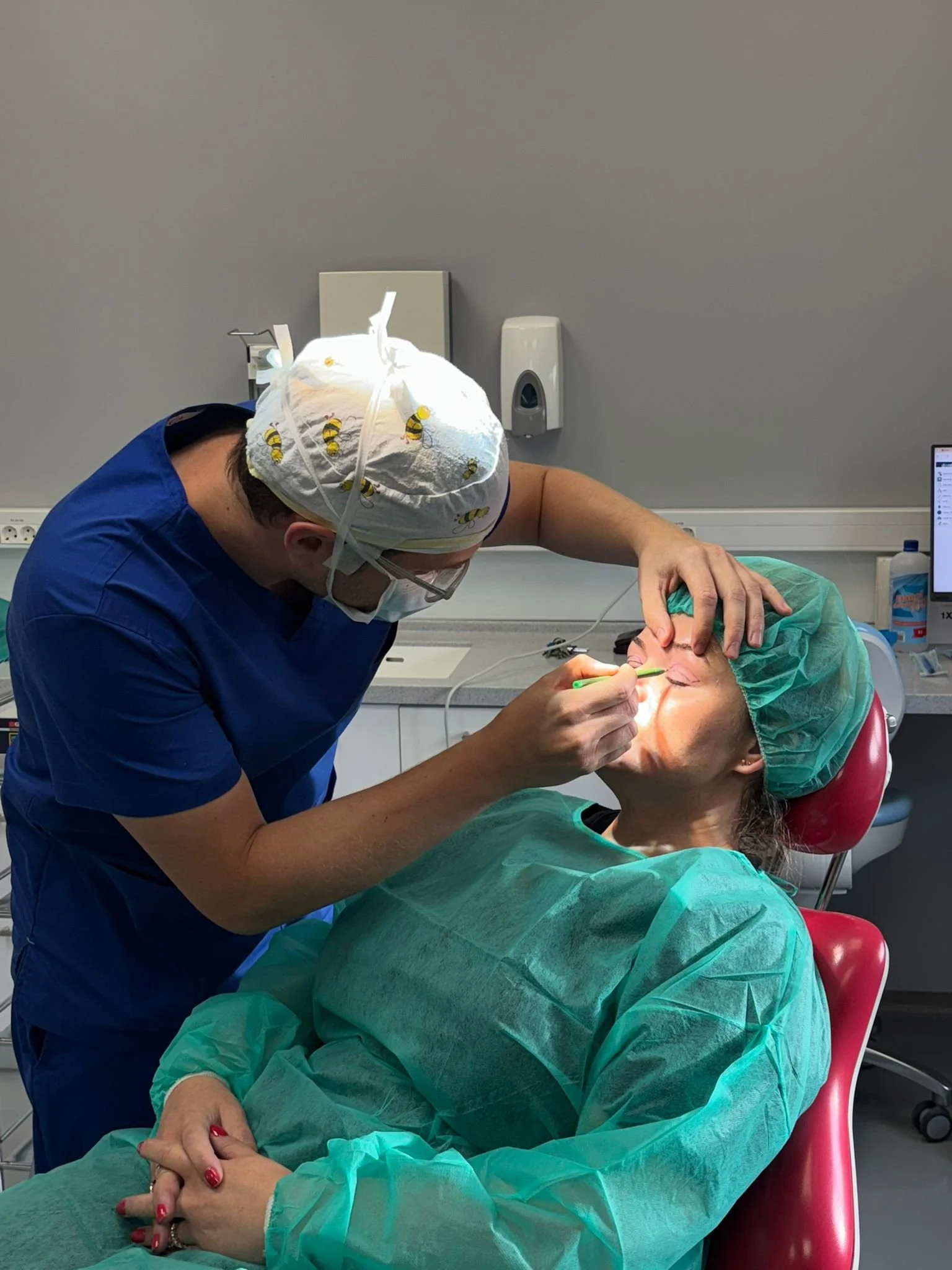 A dentist performing a dental procedure on a patient in a dental clinic. The patient is lying in a dental chair, wearing a green gown and a green surgical cap. The dentist is wearing blue scrubs, a cap with bees on it, and a mask, and is working on the patient's teeth with a tool. The clinic has medical equipment and a computer monitor in the background.