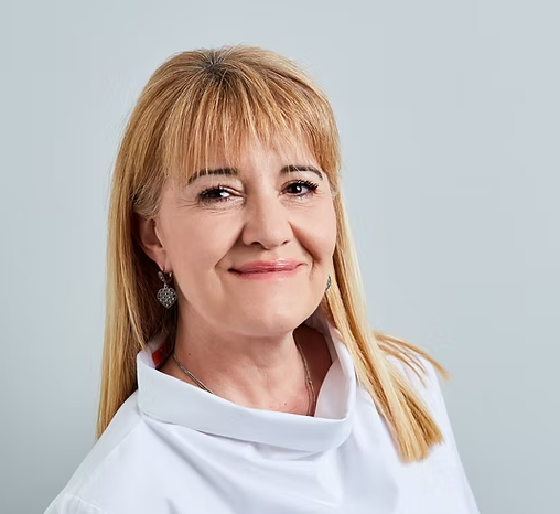 For Ars Salutaris Clinic, Portrait of middle-aged woman with blonde hair wearing earrings and a white top, smiling at the camera against a light background.