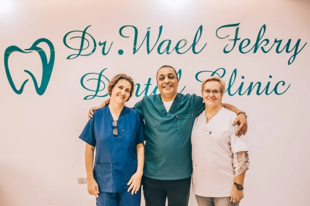 Three dental professionals standing together in front of a wall with the text 'Dr. Wael Fekry Dental Clinic' and a tooth logo. All are smiling, with the man in the middle having his arms around the women on either side.