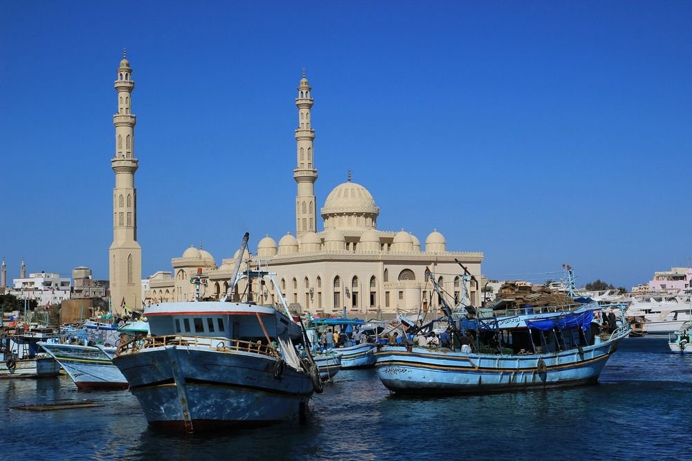 Boats docked in a harbor with a large, white mosque featuring domes and minarets in the background.