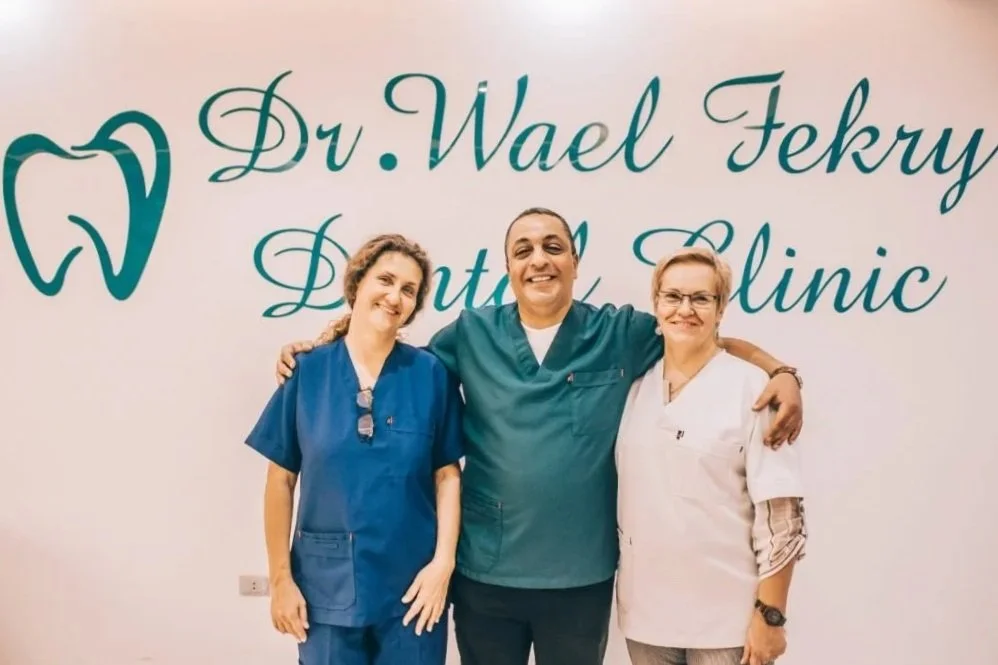 Three dental professionals standing together in front of Company Logo that reads 'Dr. Wael Fekry Dental Clinic'. The Logo has a blue tooth. The individuals are smiling and have their arms around each other, wearing dental scrubs and a white coat.