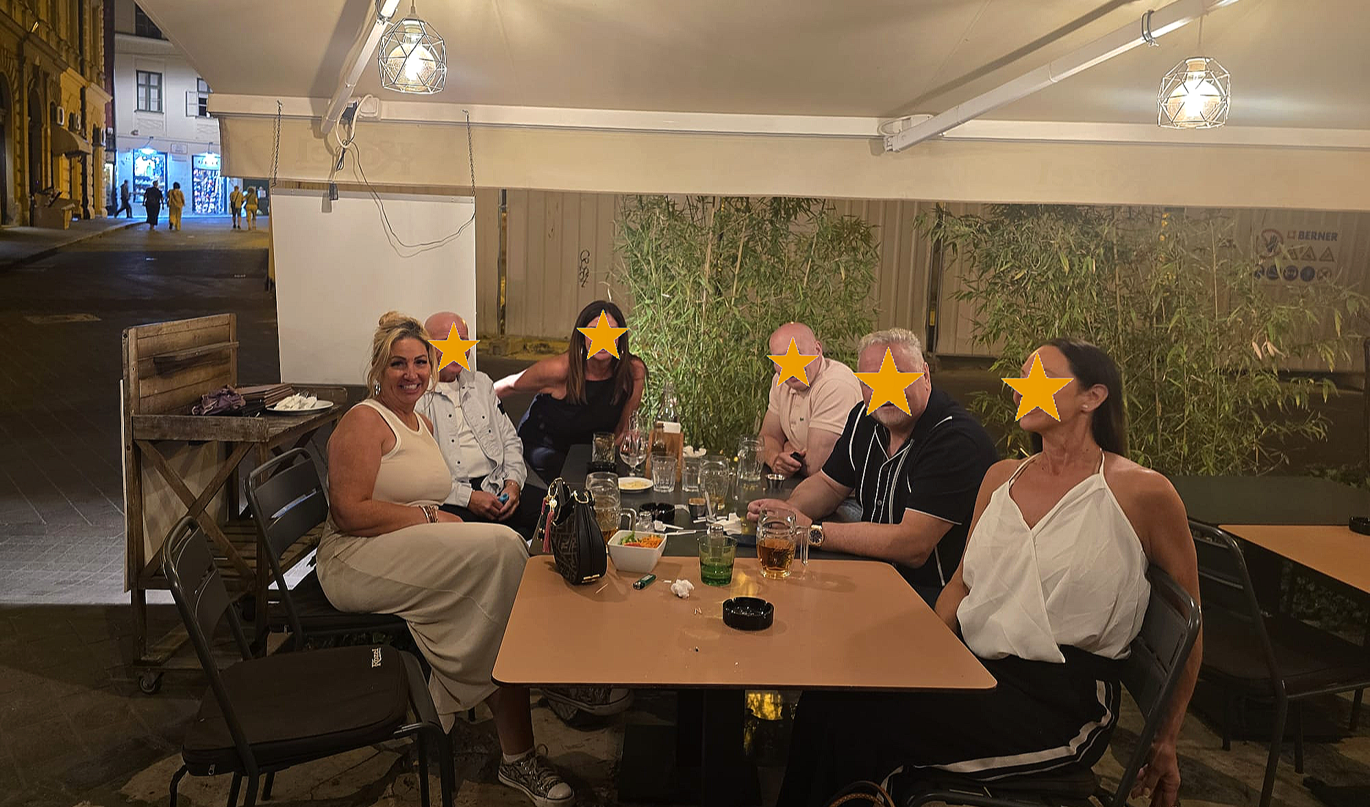 Group of seven people sitting around a table at an outdoor restaurant at night, with some laughing and smiling, and their faces obscured with star icons.