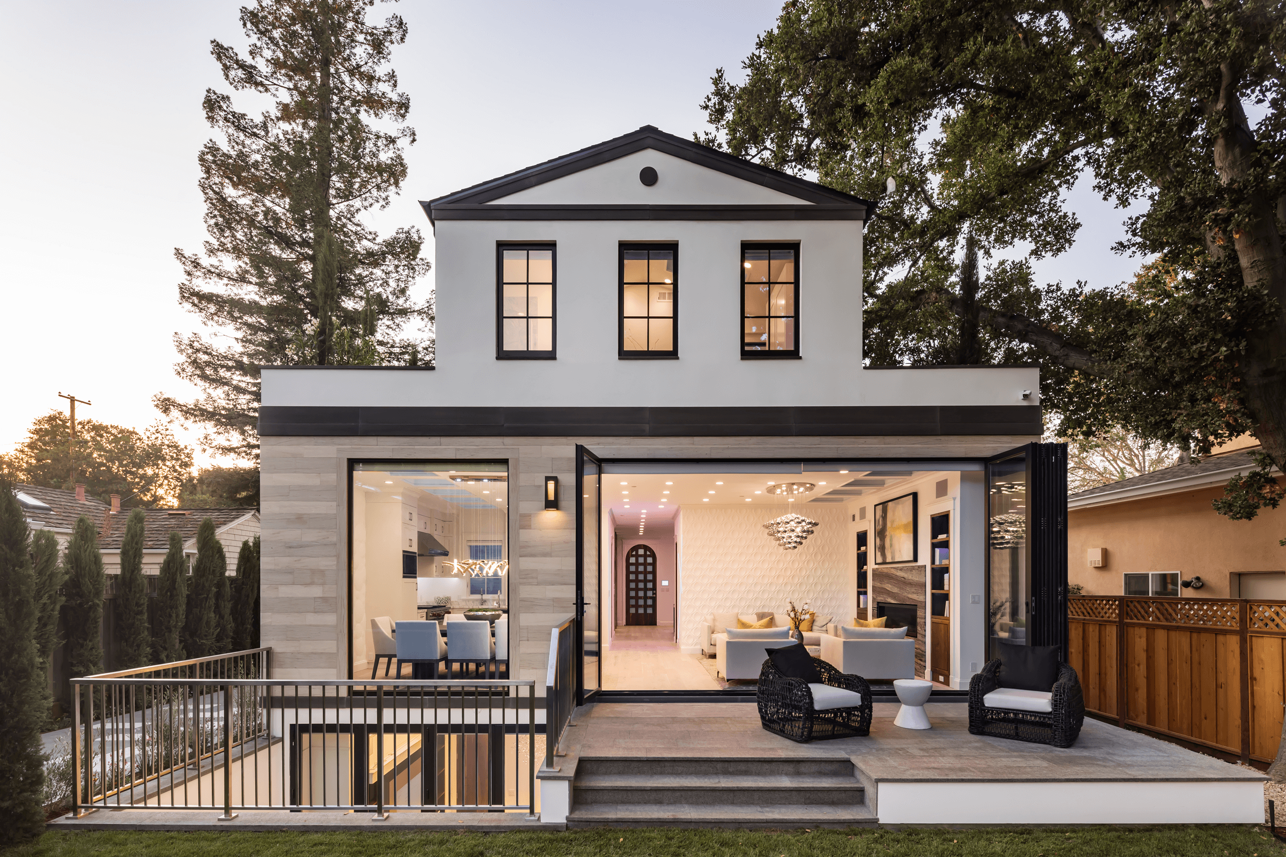 Modern two-story house with large outdoor patio area, open glass doors, and visible interior living space, surrounded by trees and a wooden fence, at dusk.