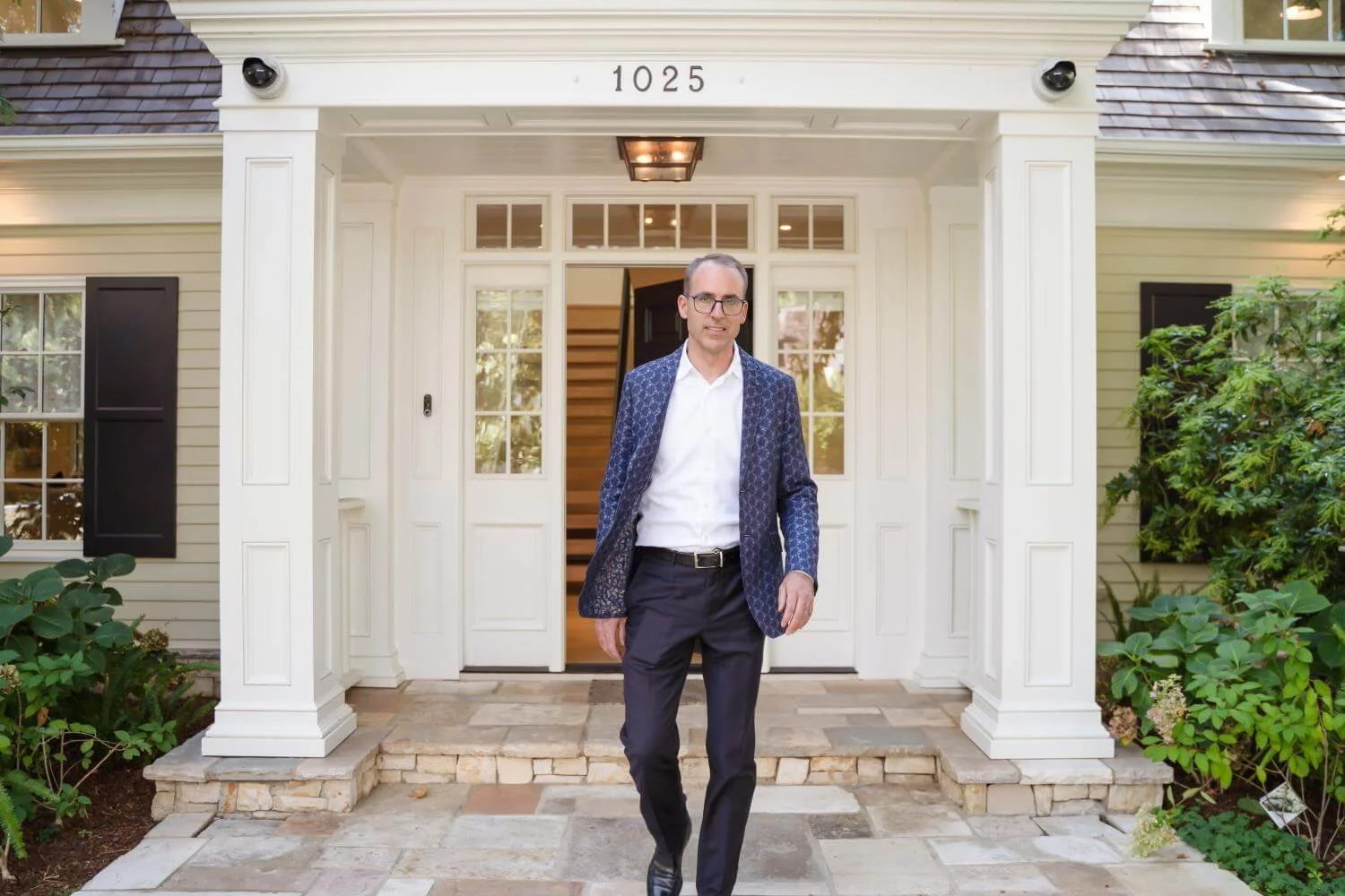 Man in a patterned navy blazer, white shirt, and glasses walking out of a white house with black shutters, green plants, and stone steps.
