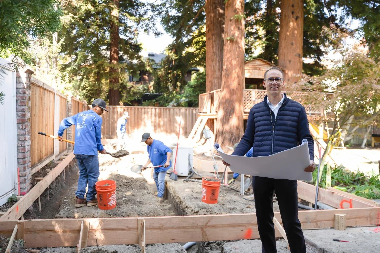 Man holding blueprints standing in front of a construction site for a backyard deck, with workers shoveling dirt and working on the ground, surrounded by large trees and a wooden fence.