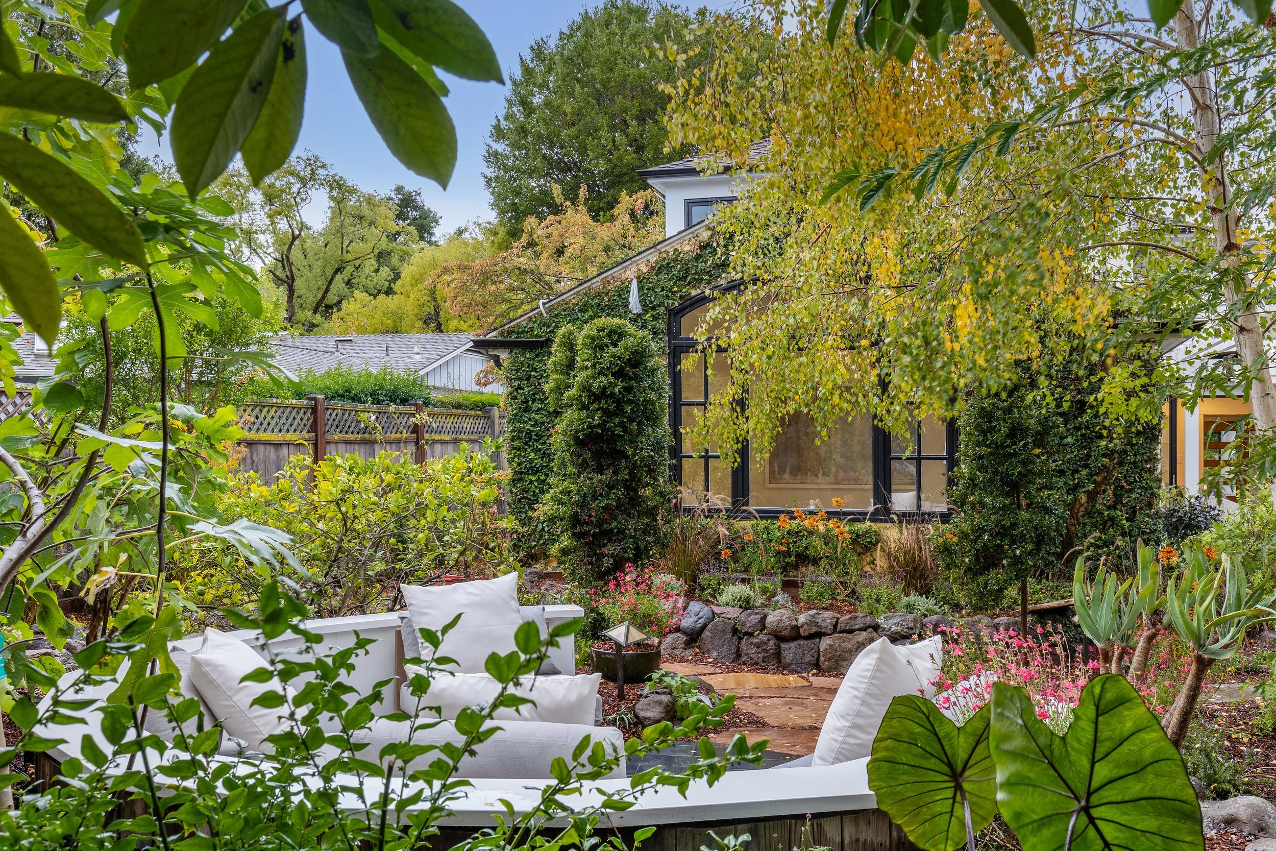 A backyard garden with lush green trees and shrubs, colorful flowers, a cozy white outdoor seating area with cushions, and a house with large windows partially covered by climbing plants.
