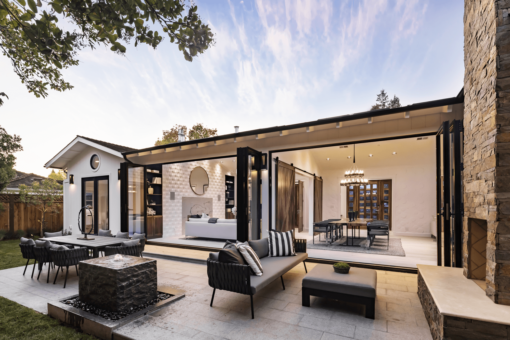 A modern house with an open living space, outdoor patio furniture, and a view of a blue sky with wispy clouds, surrounded by trees and a wooden fence.