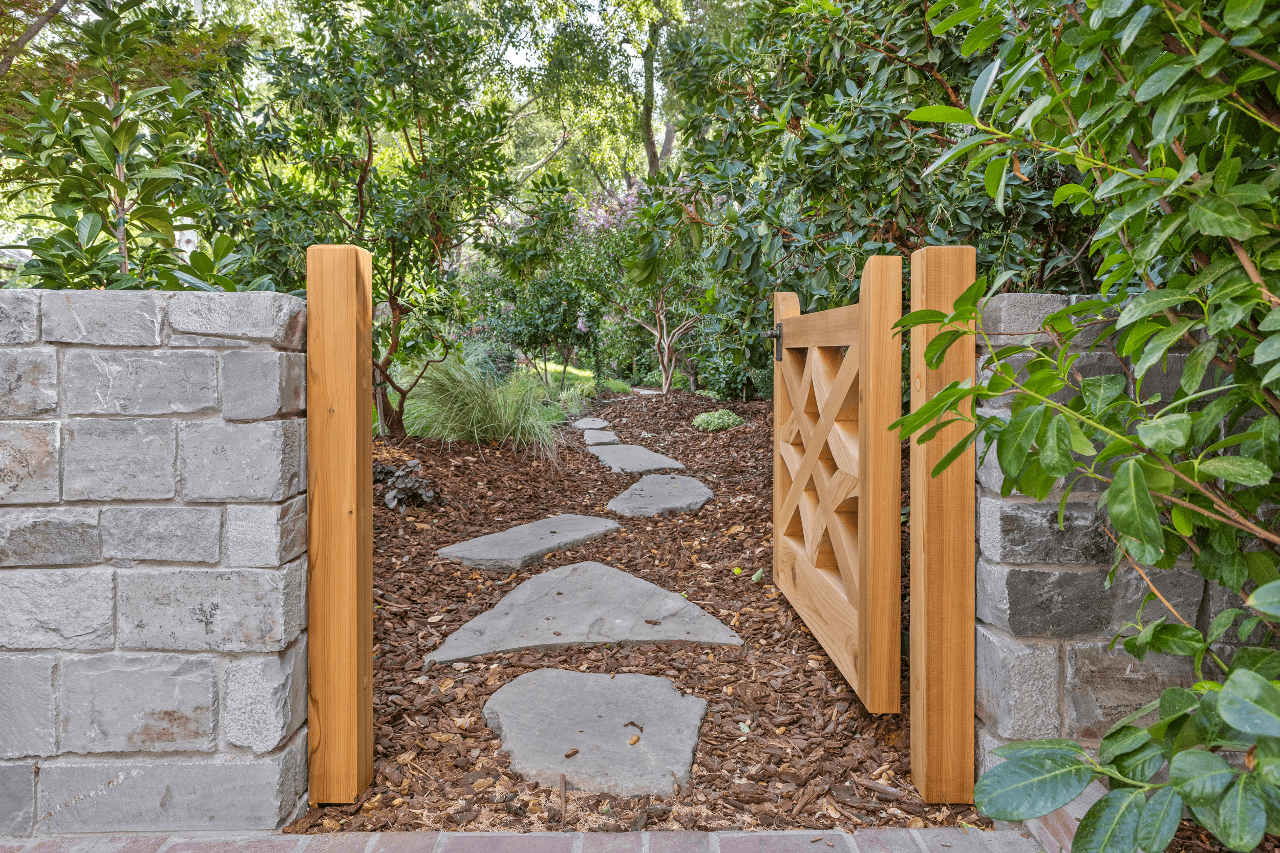 A garden pathway with stone stepping stones, surrounded by greenery and bushes, with a wooden gate and brick walls on each side.