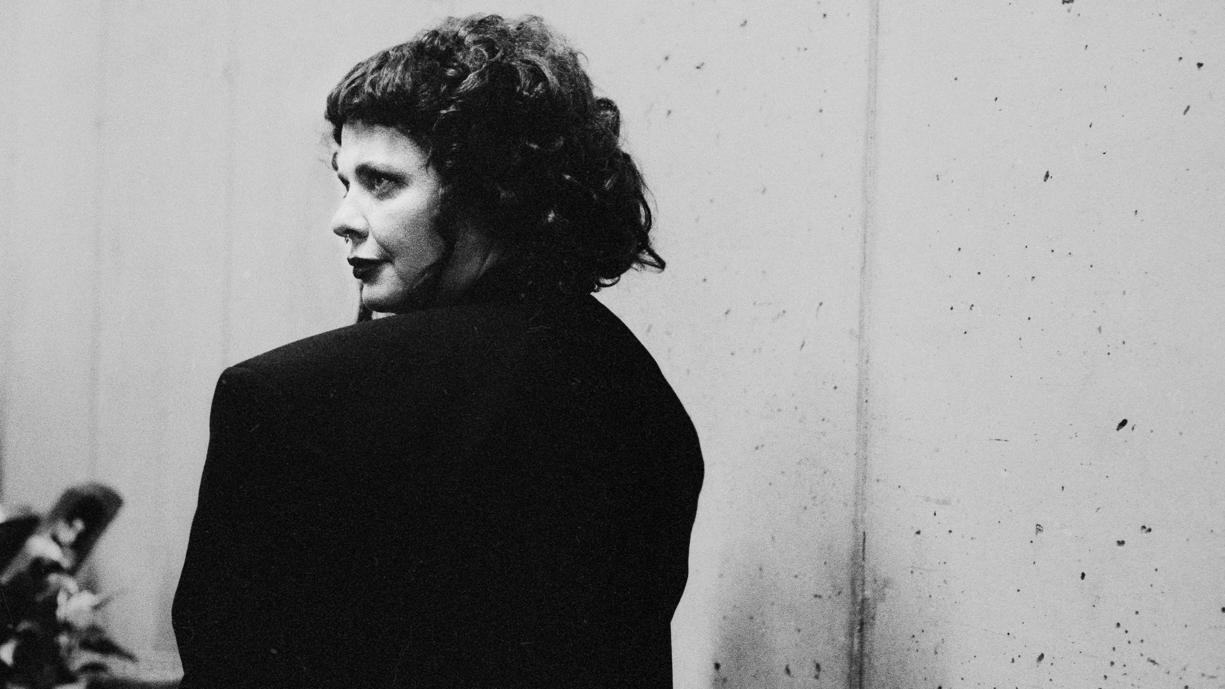 A woman with curly hair and dark lipstick stands sideways against a plain wall, looking over her shoulder at the camera.