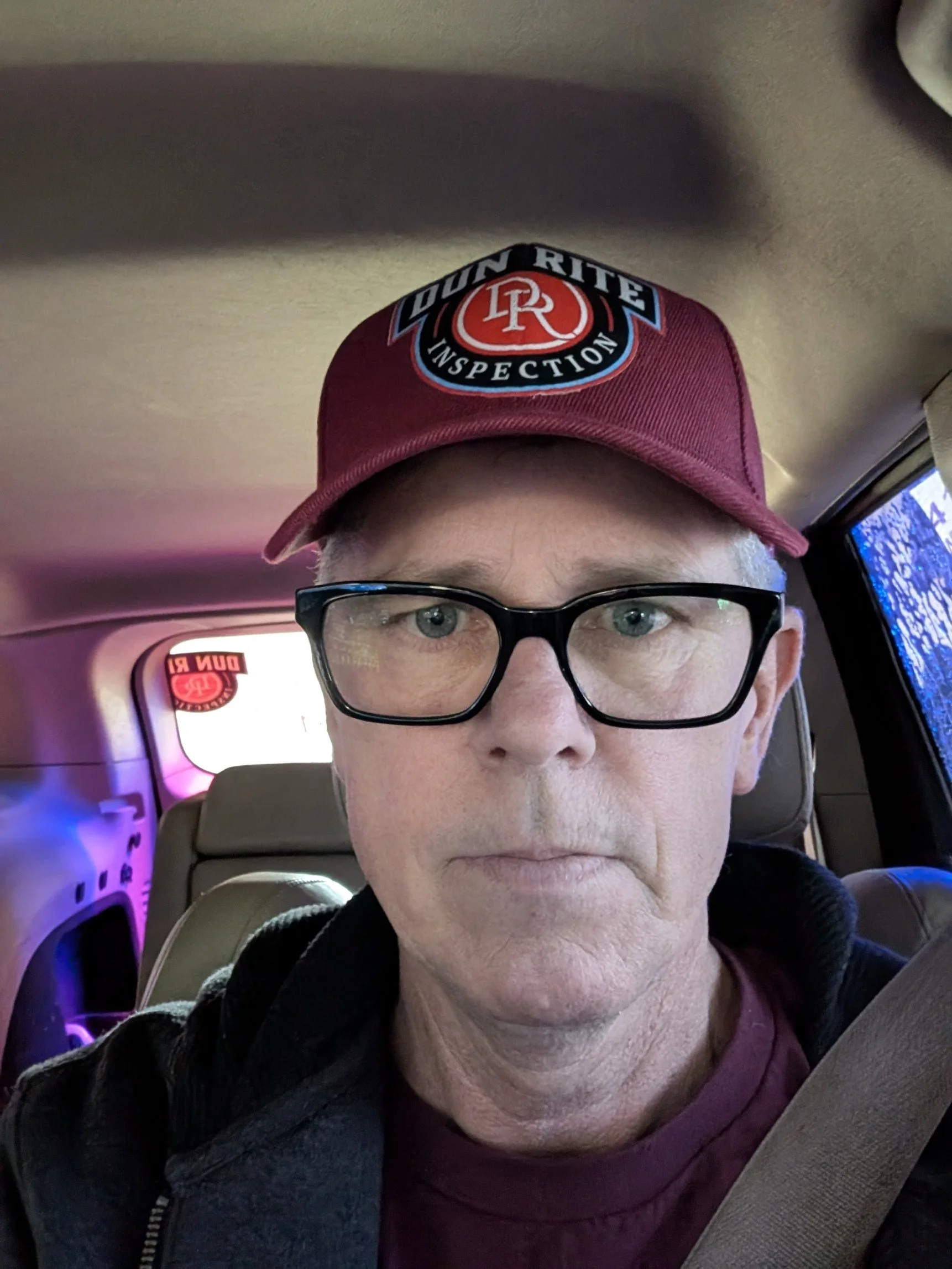 A man wearing glasses and a maroon cap with the logo 'DuN RITE INSPECTION' is sitting inside a vehicle, with a seatbelt across his chest. The vehicle's interior is beige, and there are some colorful lights and screens in the background.