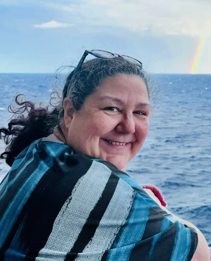 A smiling woman with curly hair and glasses resting on her head, sitting outdoors on a boat or near the ocean with a rainbow in the background.
