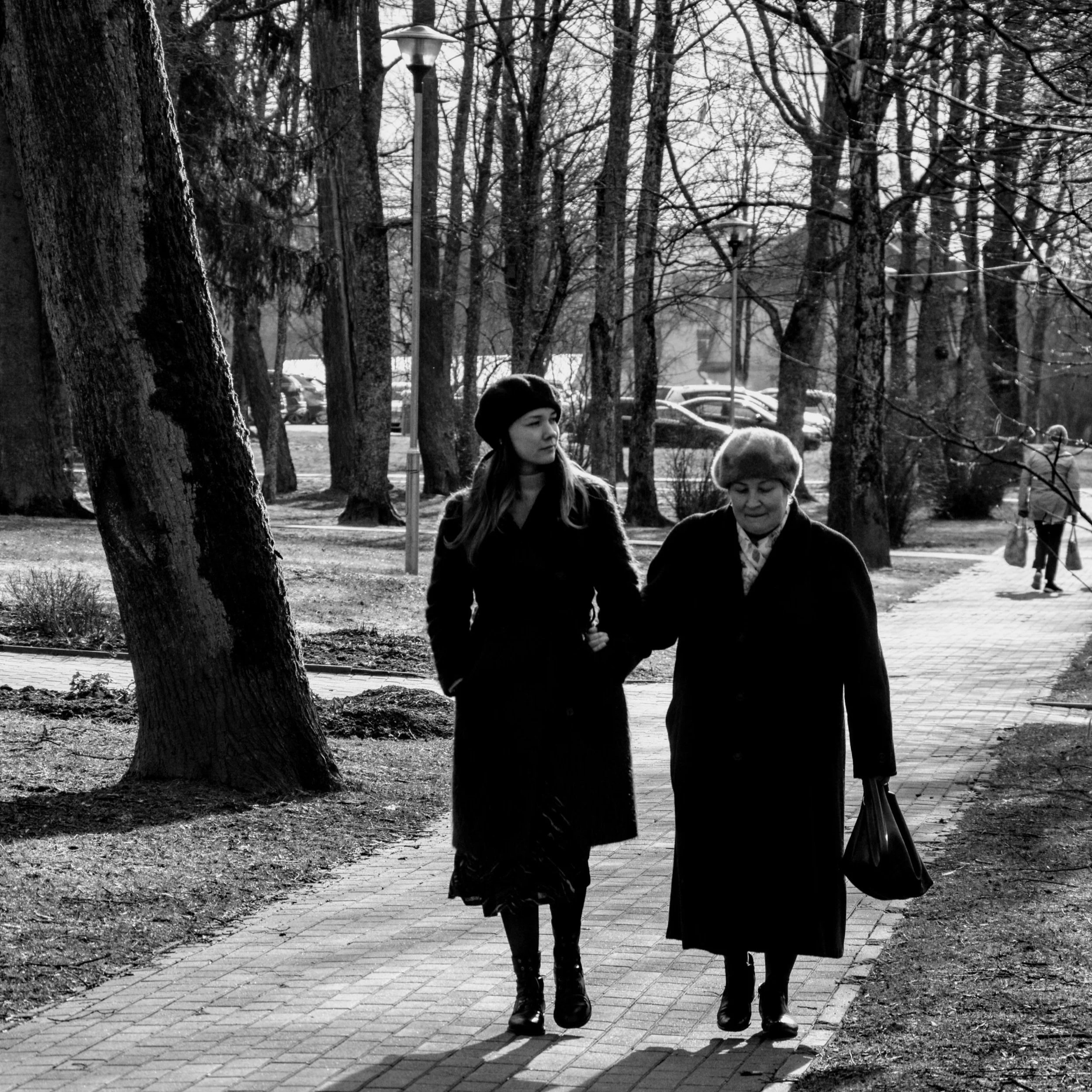Two women walking together on a paved path in a park with tall trees, one younger and one elderly, both dressed warmly and carrying bags, with parked cars and a person in the background.