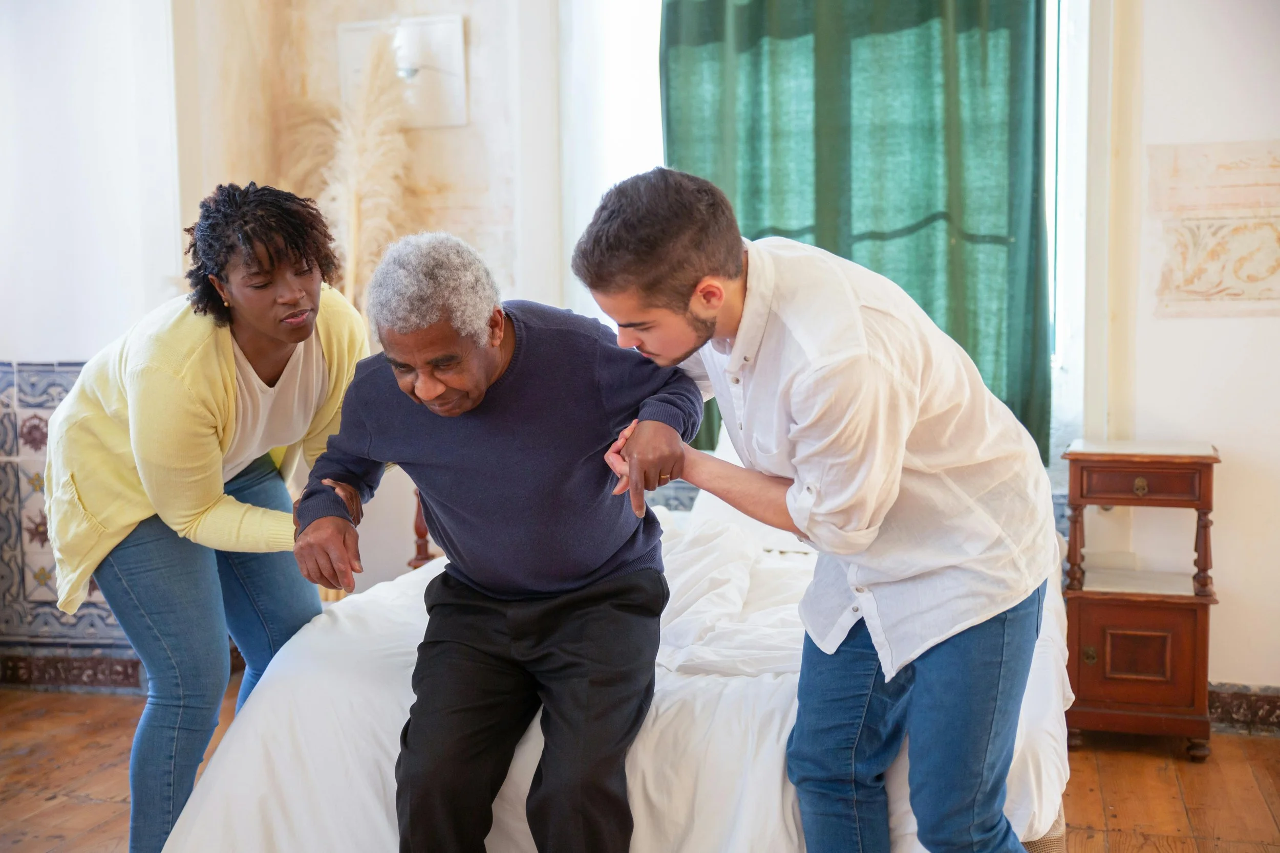 A young man and a young woman help an elderly man stand up from a bed in a brightly lit room, with a woman watching supportively.