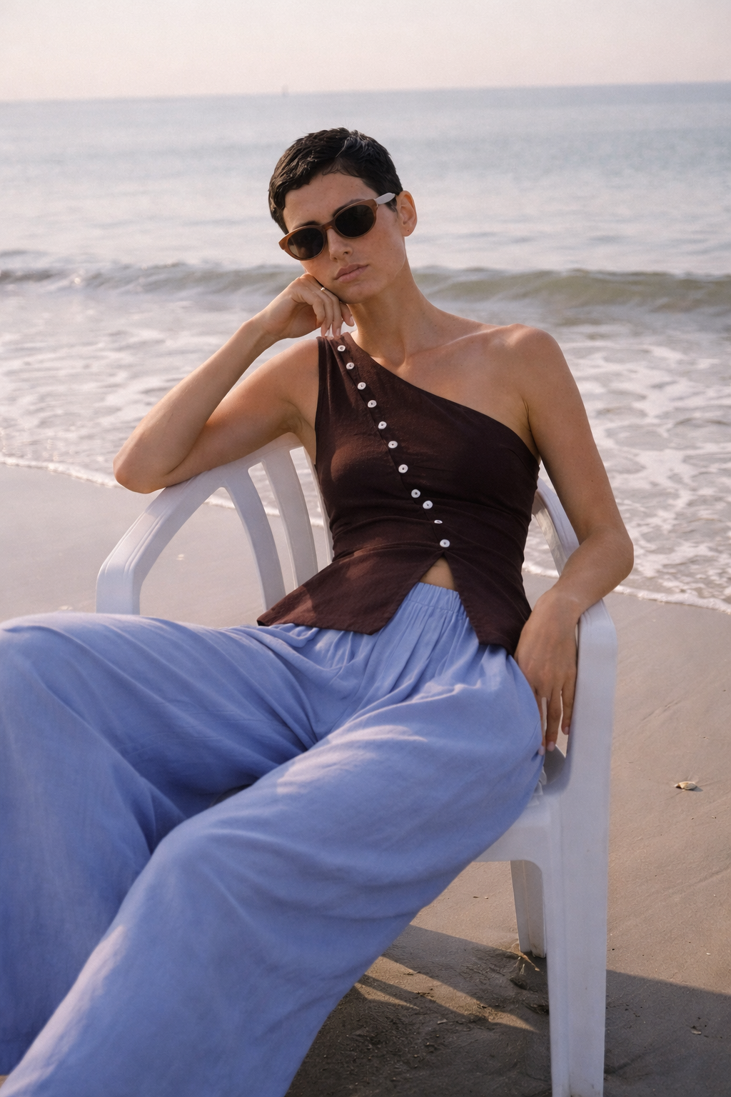 woman sitting down on white plastic chair on beach wearing coffee colored top with diagonal button line