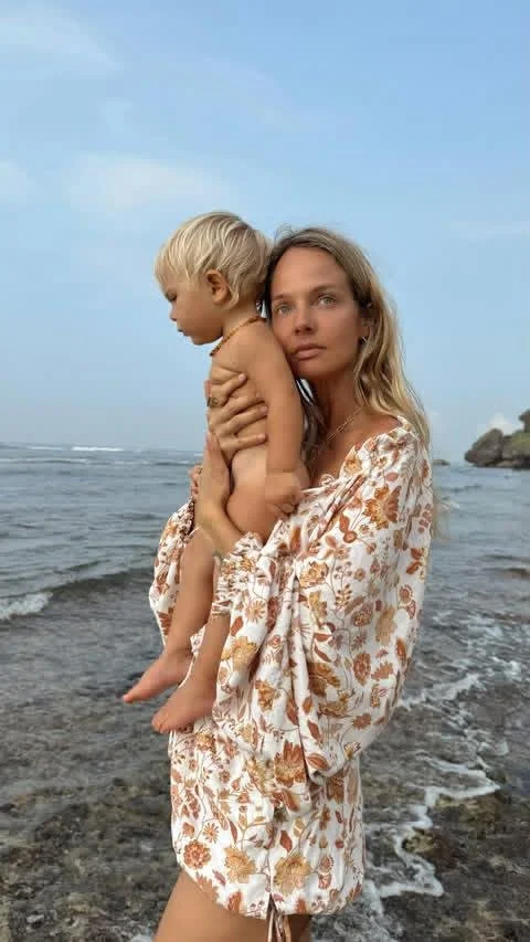 A woman holding a young boy at the beach, with ocean waves and a partly cloudy sky in the background wearing Avery Jumpsuit of Sutura Studio 
