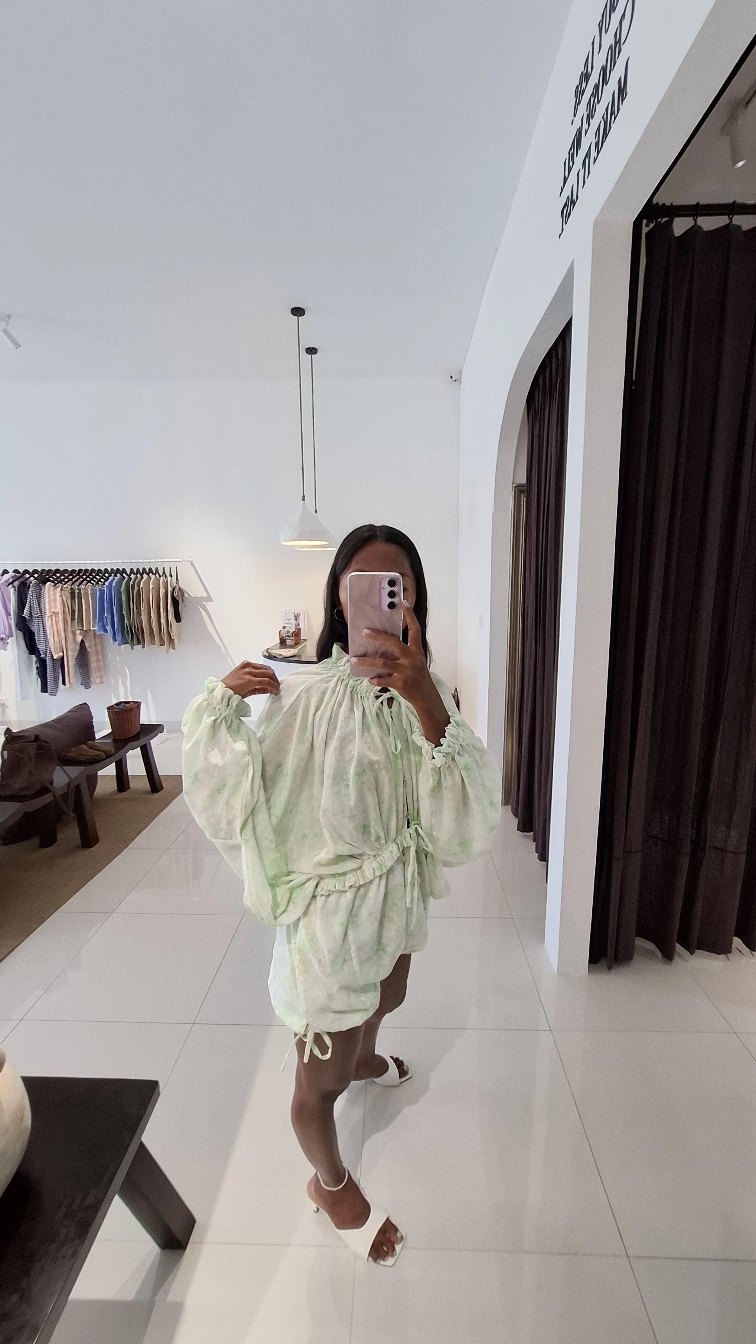 A woman taking a mirror selfie in a fitting room, wearing a light-colored, tie-dye Avery Jumpsuit with puffed sleeves and ruffled details from Sutura Studio, paired with white high-heeled sandals.