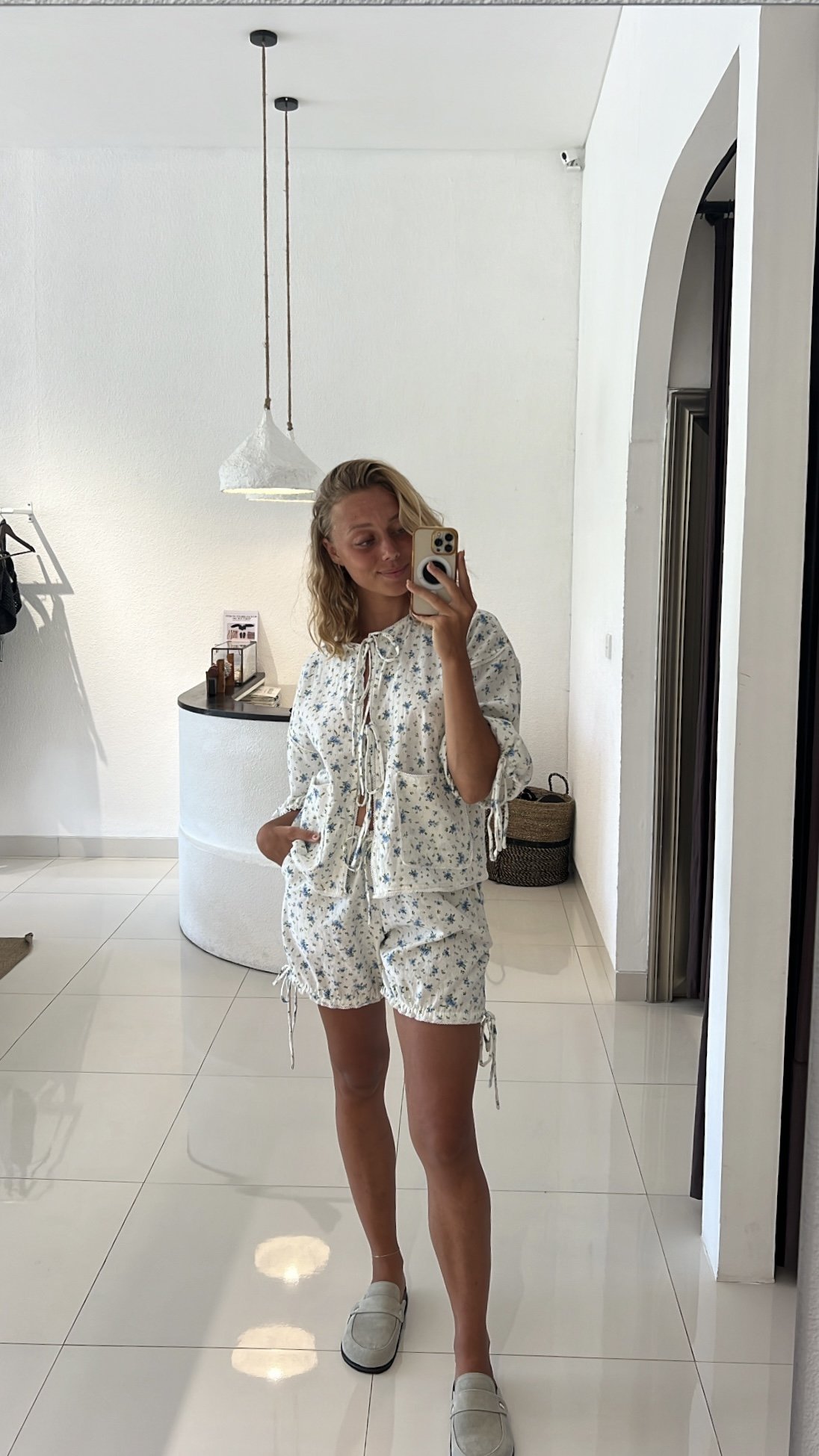 A woman taking a mirror selfie in a minimalist, white-tiled room. She is wearing a cotton set from Sutura Studio with a floral pattern and loafers, and is smiling softly.