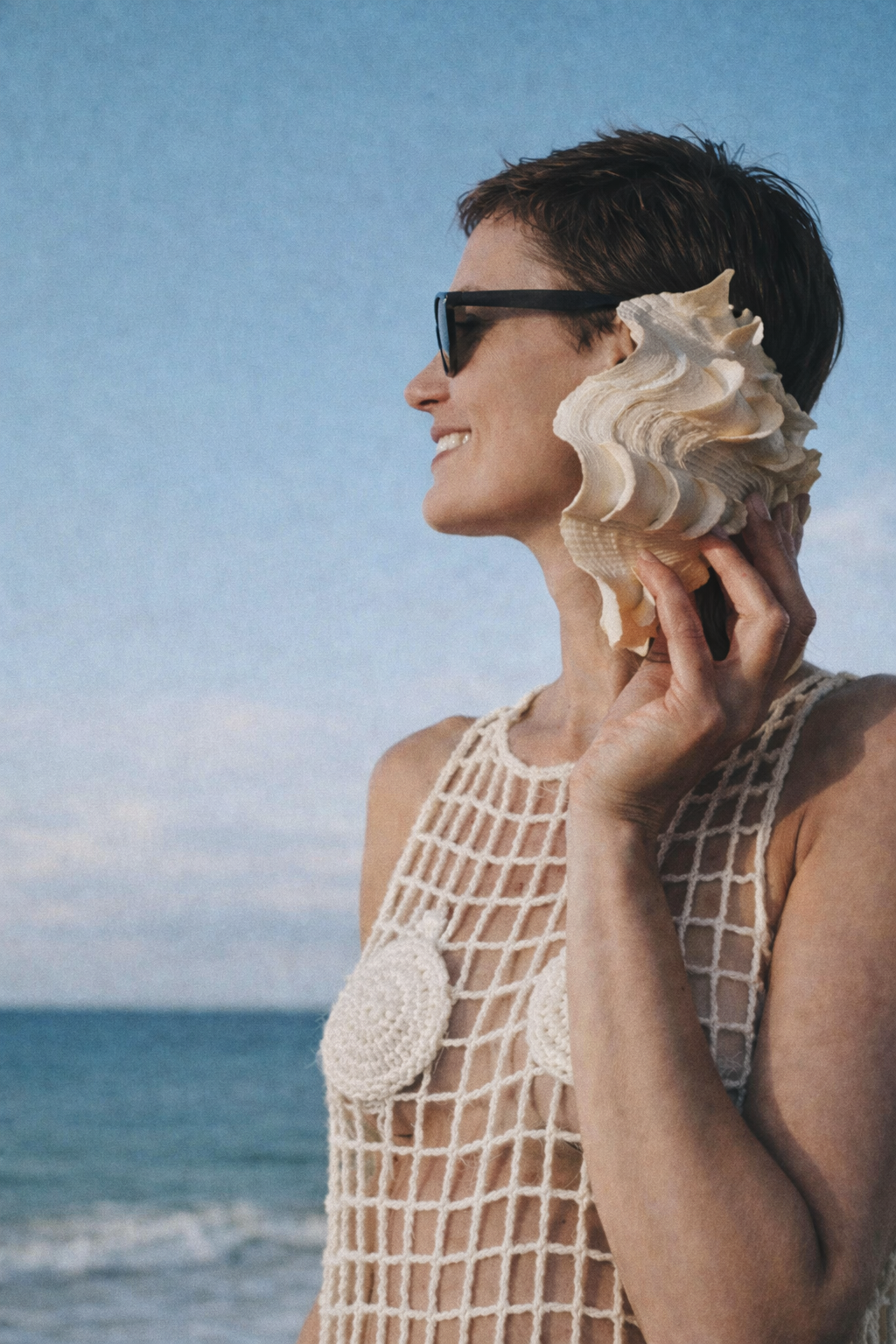 woman holding a large shell next to her face wearing a sunglasses standing on a beach wearing a crochet top with crochet shell detail on breasts