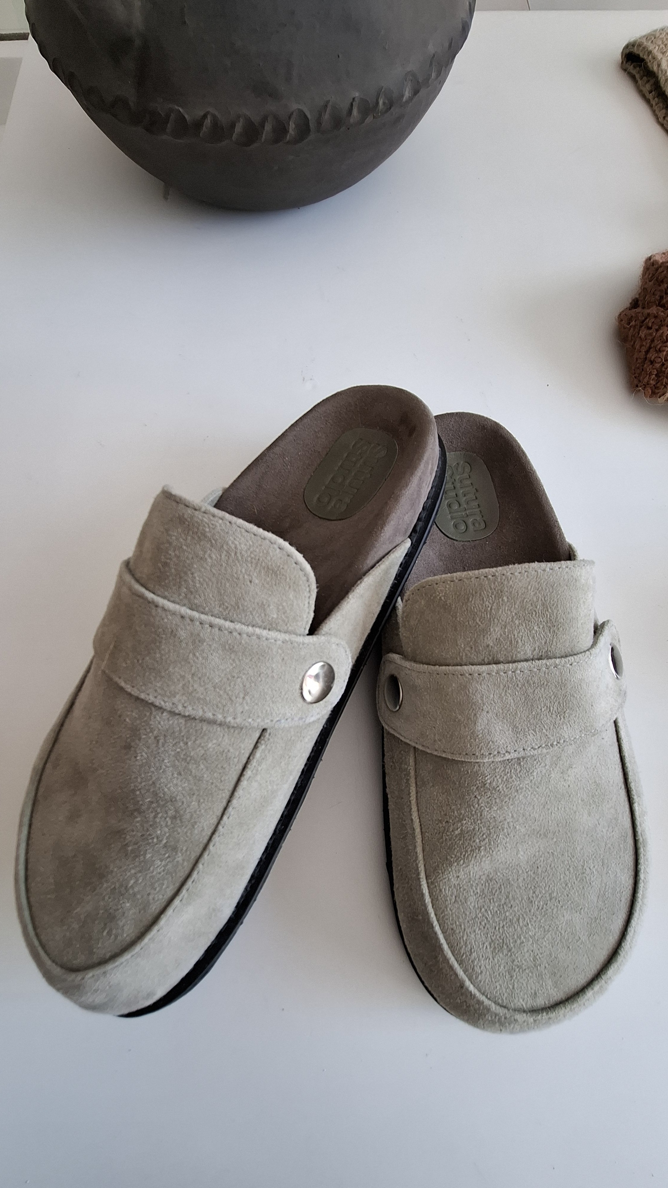 A pair of beige suede Sutura clog-style slippers placed on a white surface.