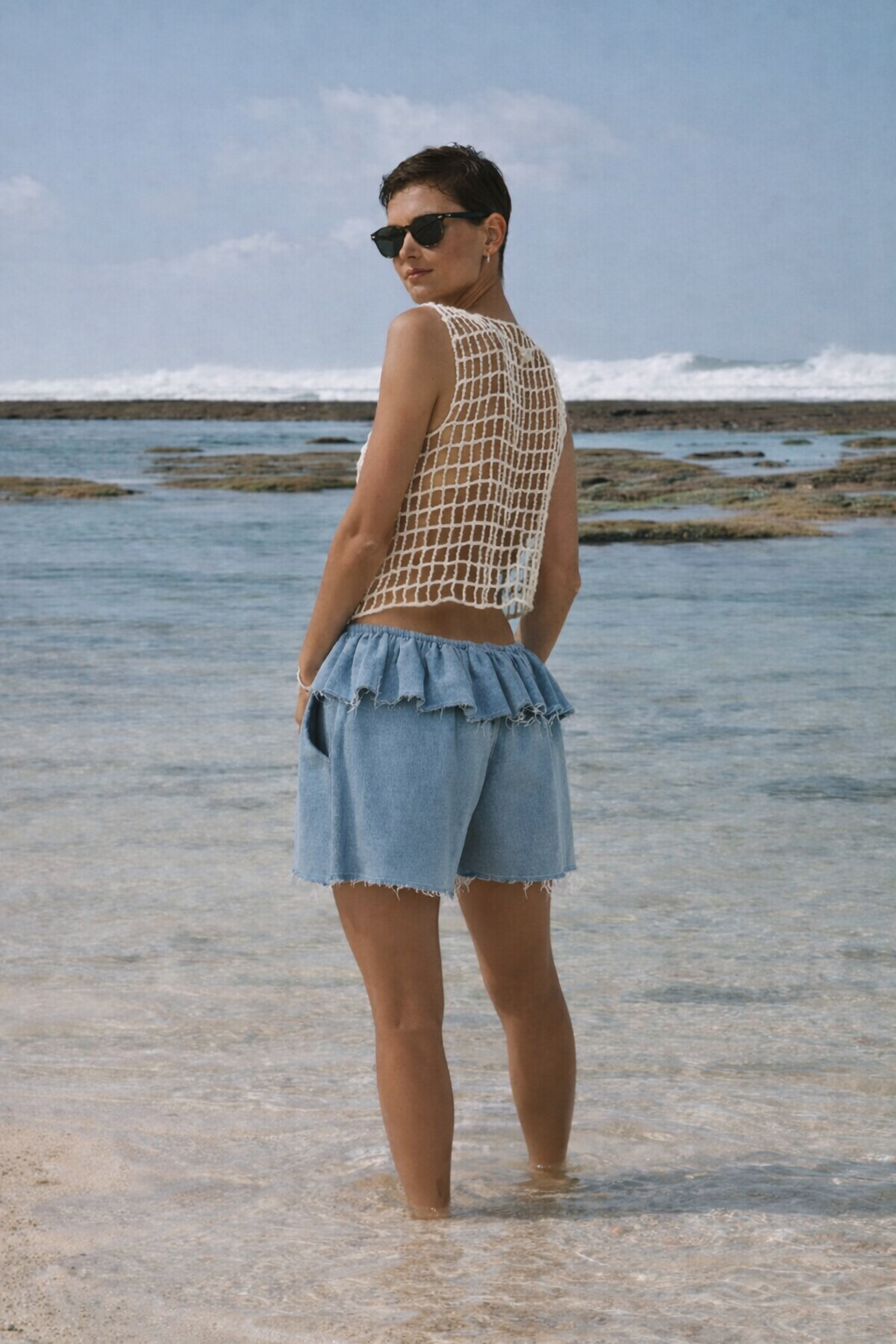 woman standing on beach with her back, turning her face, wearing Renata denim shorts and crochet top