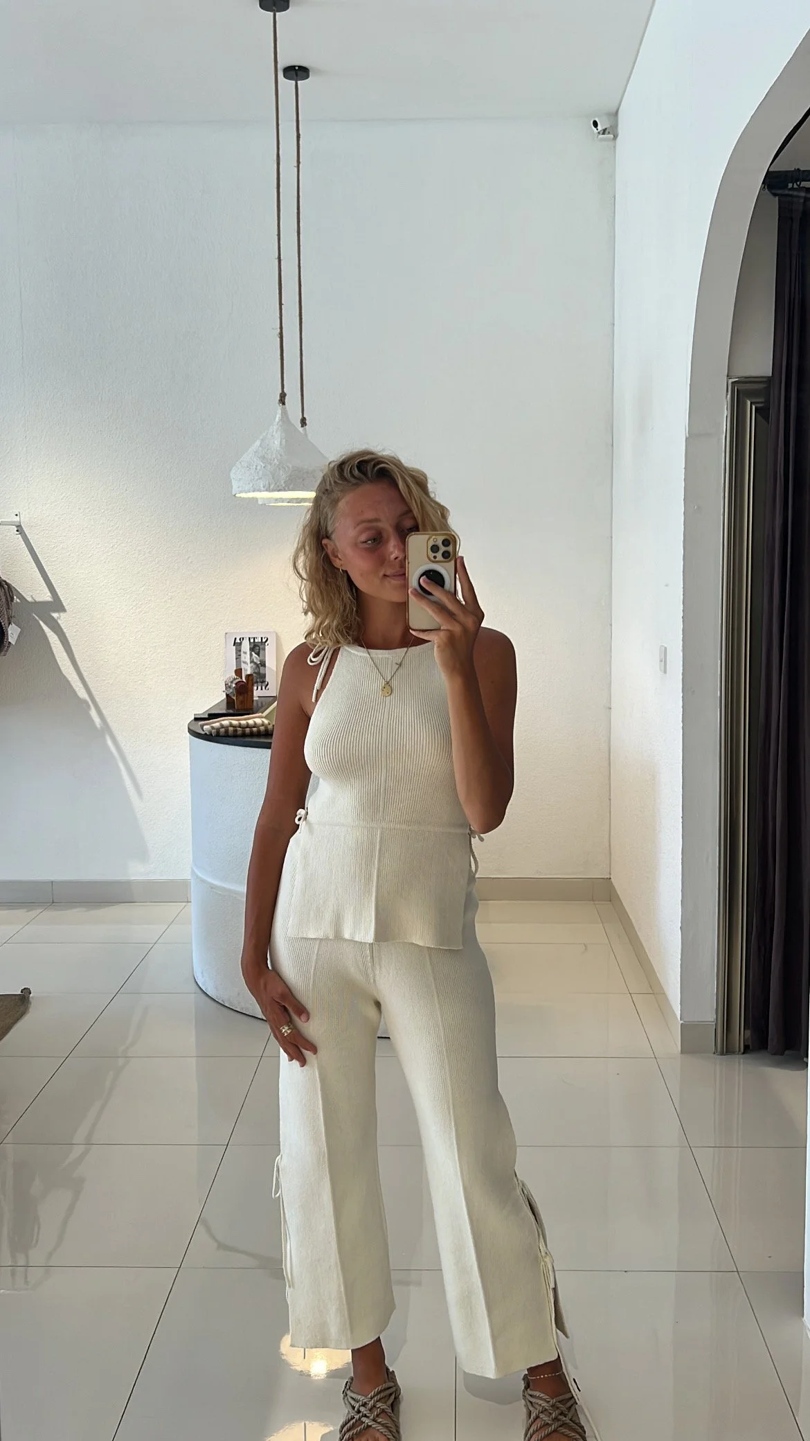 A woman taking a mirror selfie in a minimalistic room wearing a cream-colored sleeveless top and matching wide-leg pants from Sutura Studio with wavy blonde hair and some jewelry.