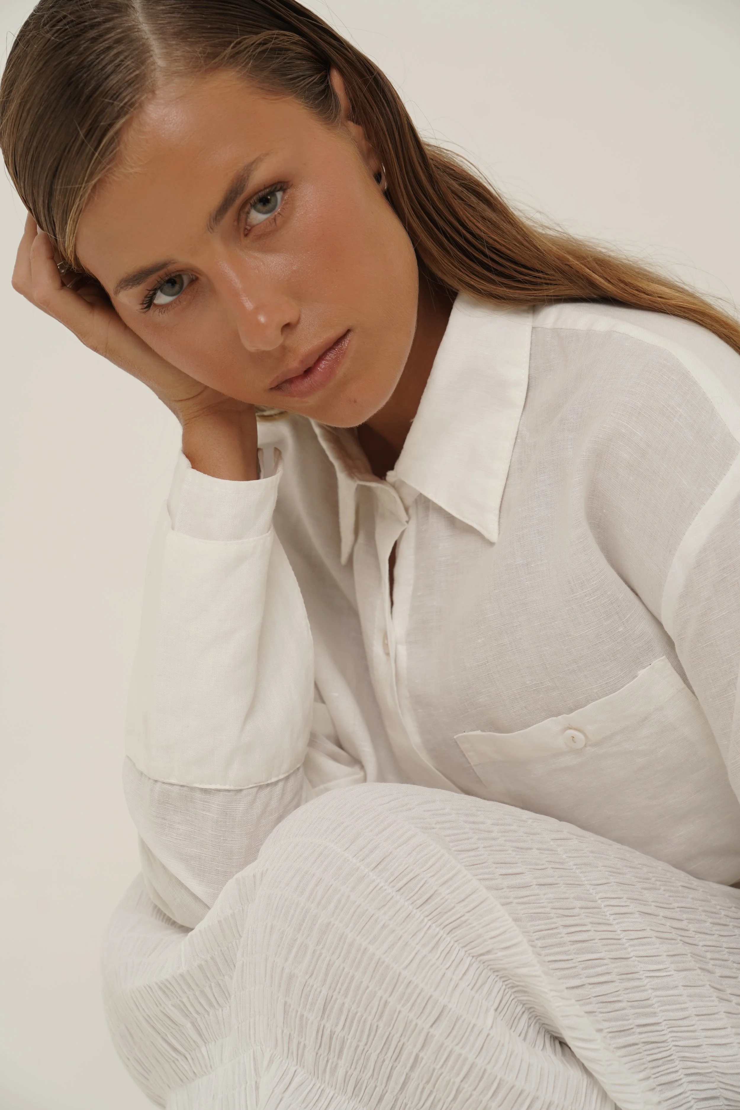 Woman sitting down wearing Reformation Shirt in white linen holding her face