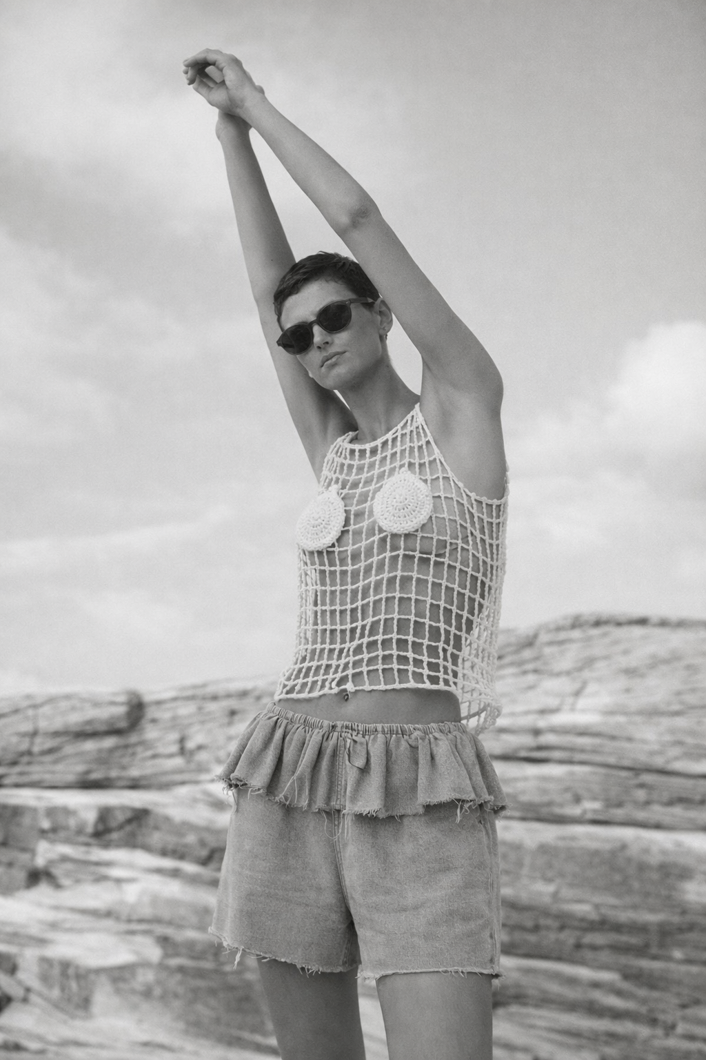 woman standing on beach with crochet top and denim shorts, stretching her hands in black and white