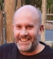 A smiling man with a beard and shaved head outdoors, with wooden logs and a fence in the background.