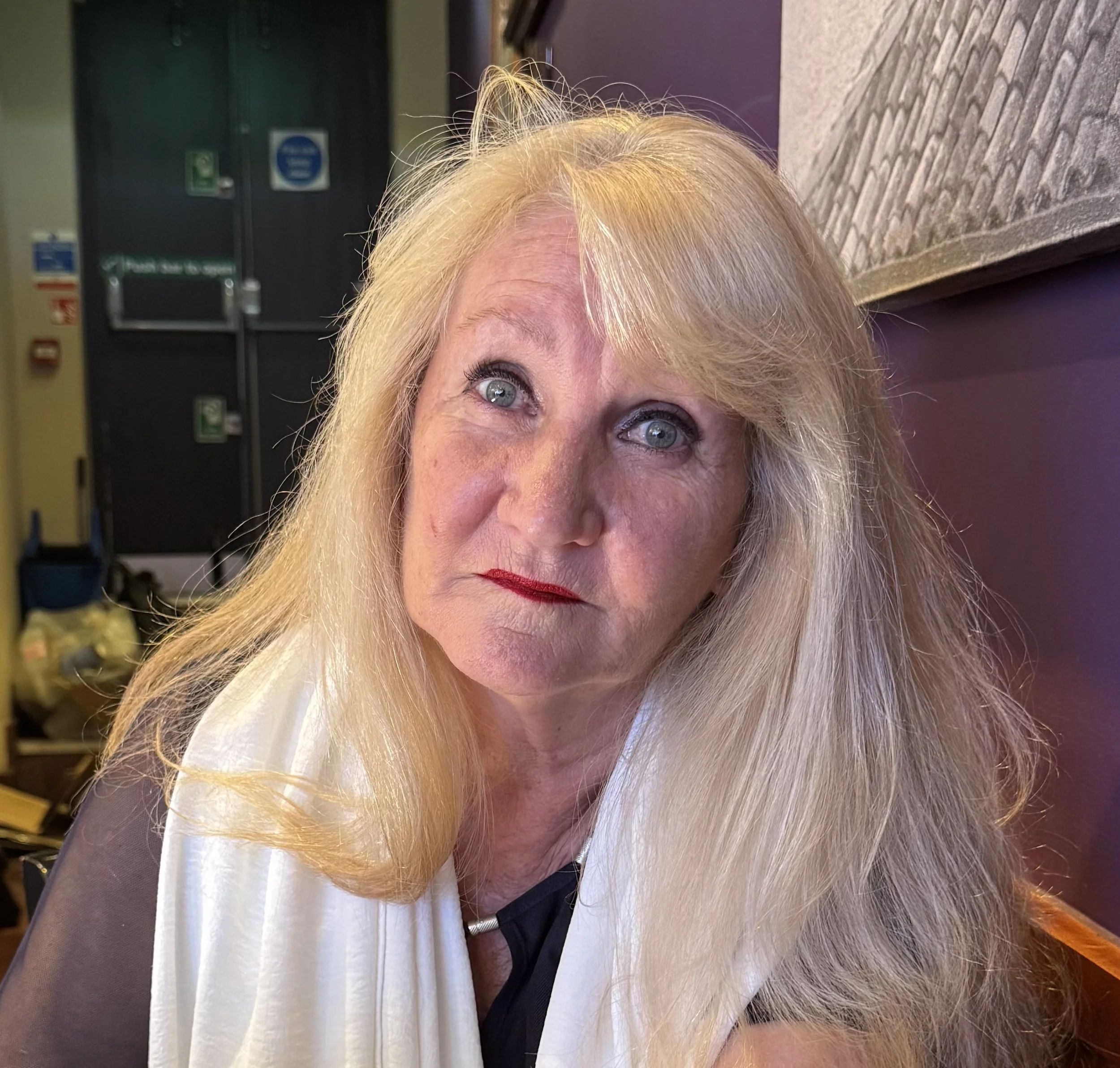 Close-up portrait of a blonde woman with blue eyes, red lipstick, wearing a black top and a cream-colored scarf, sitting indoors against a purple wall with framed art and a door in the background.