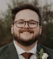 A smiling man with glasses and a beard, wearing a suit and tie, outdoors with trees in the background.