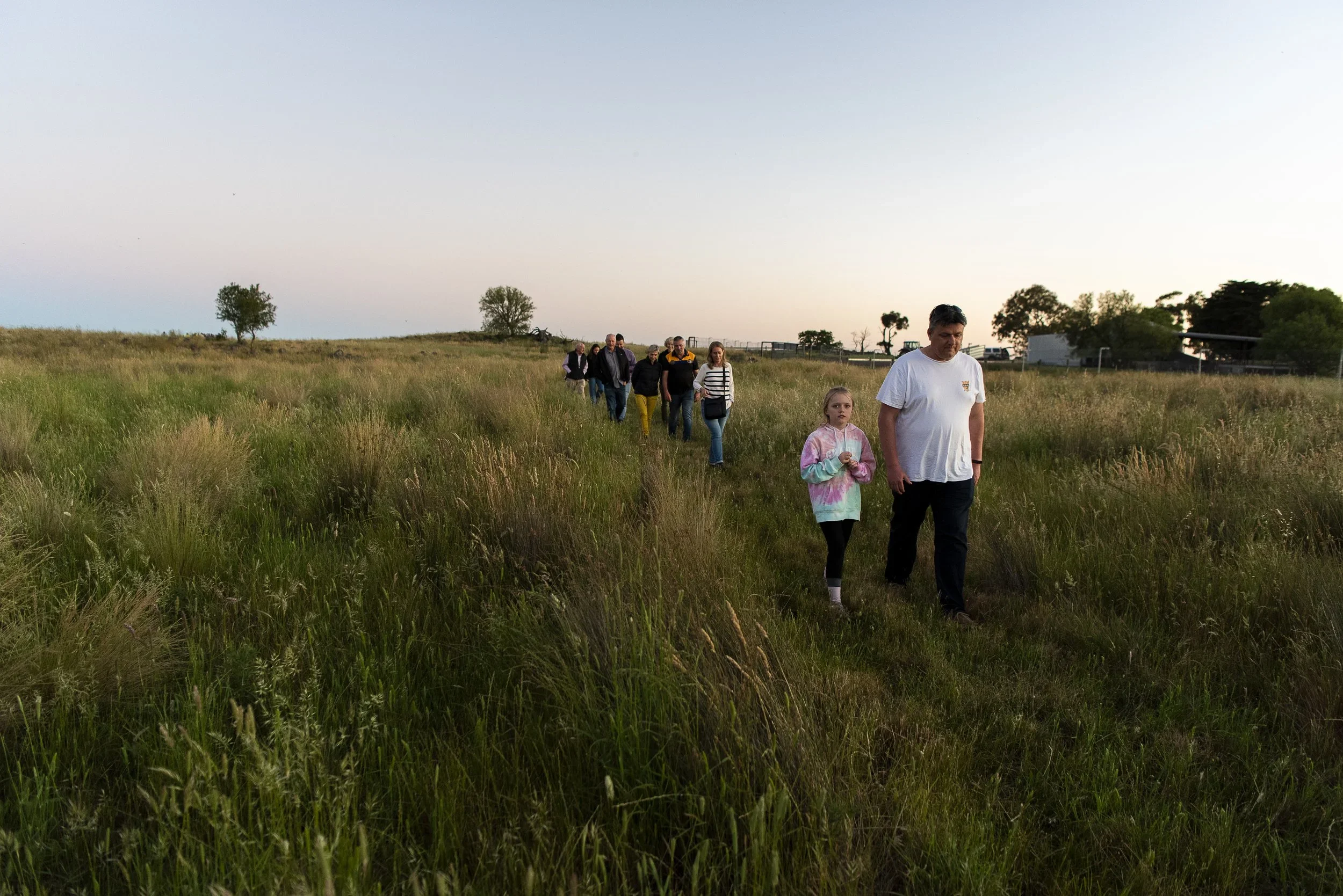 Group of people walking in a grassy field at sunset, with a clear sky and some trees in the background.