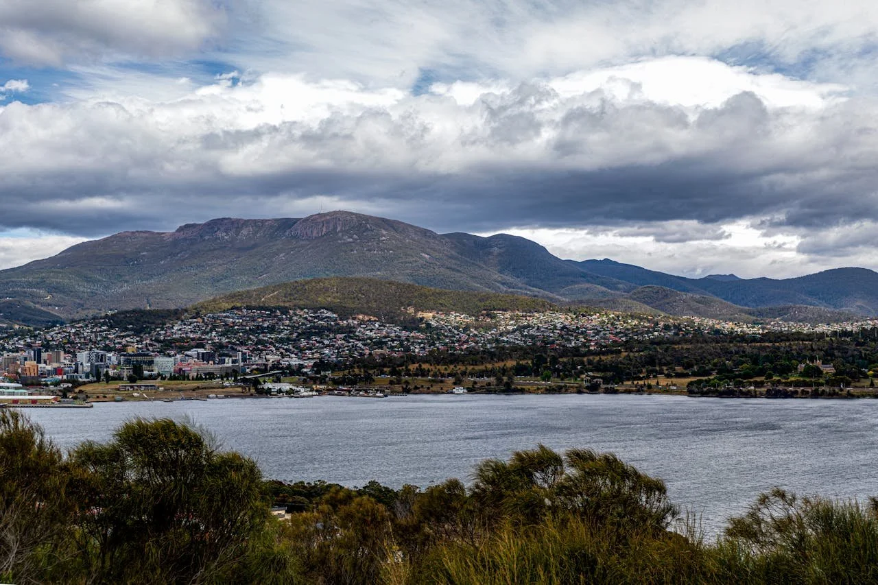 A scenic view of a large body of water in the foreground, with trees along the shore, and a city nestled at the base of a mountainous landscape under a cloudy sky.