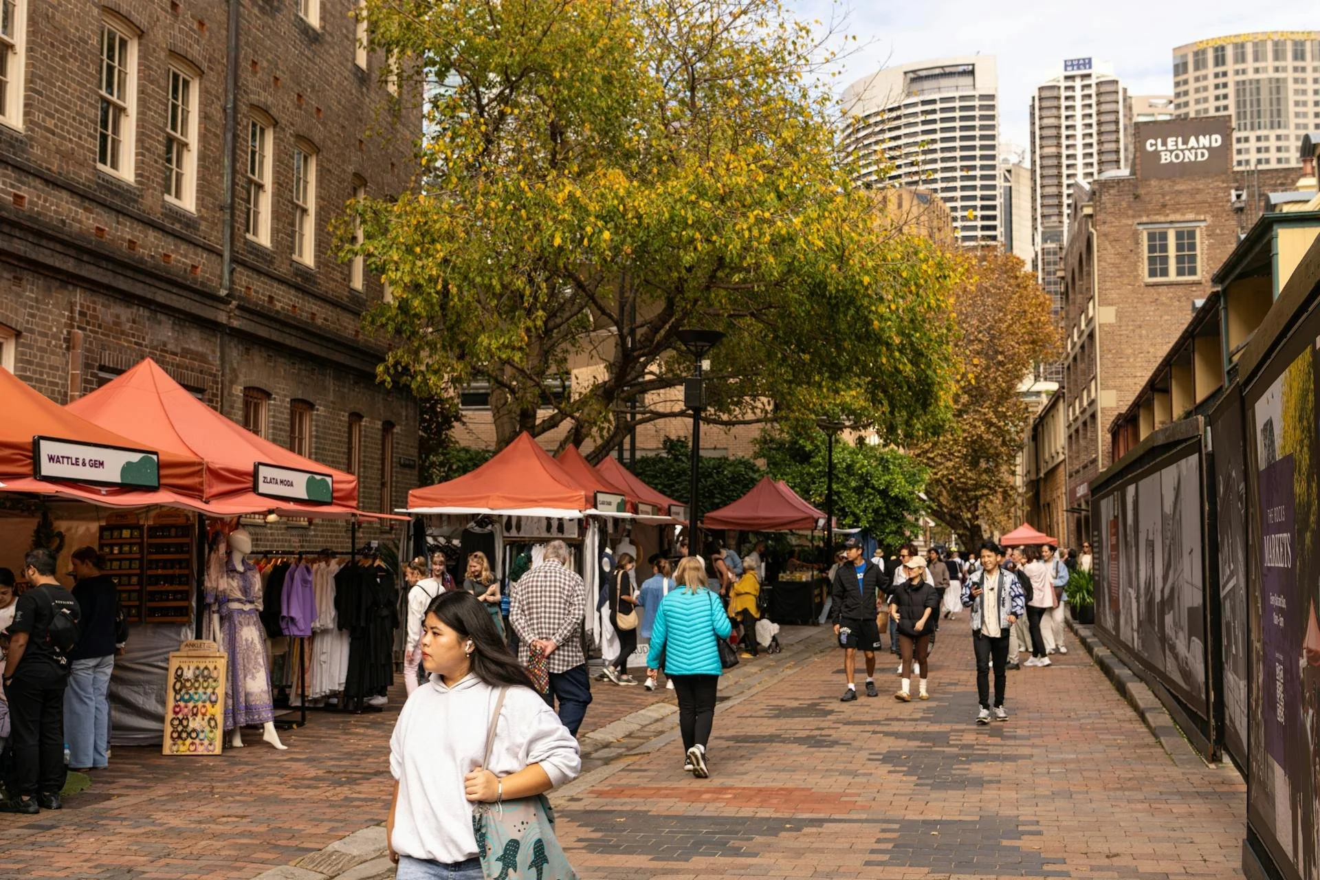 Outdoor street market with orange tents, people shopping and walking, brick pavement, trees with autumn leaves, and tall buildings in the background.