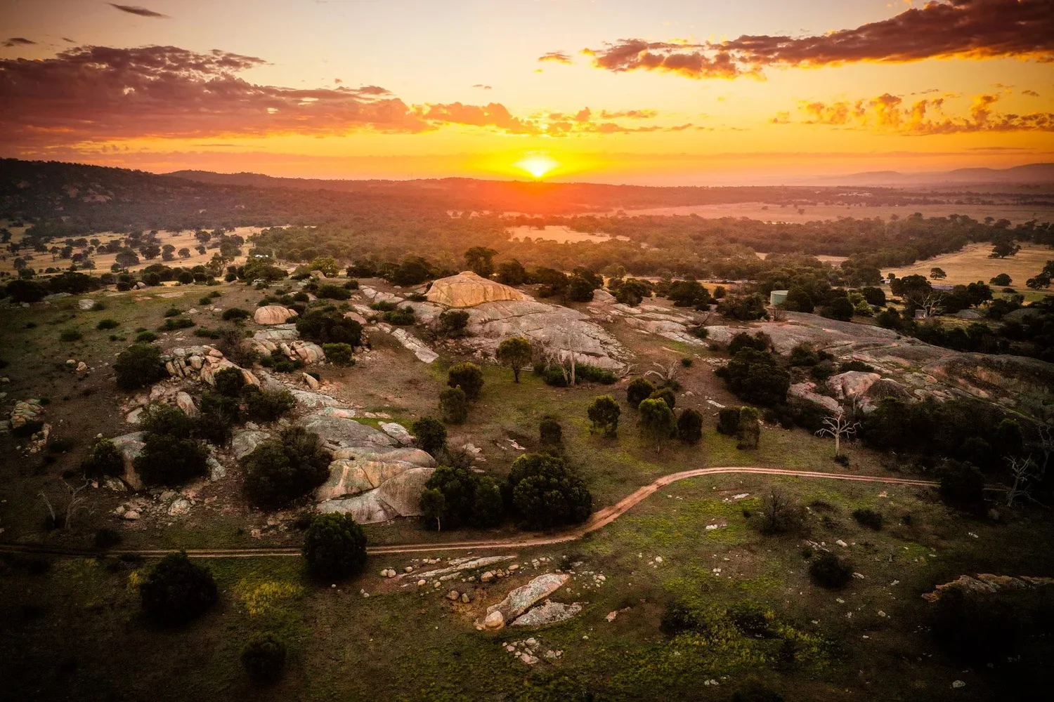 A landscape of rocky terrain with scattered trees under a colorful sunset sky. A dirt path winds through the scene, with mountains and a distant horizon.