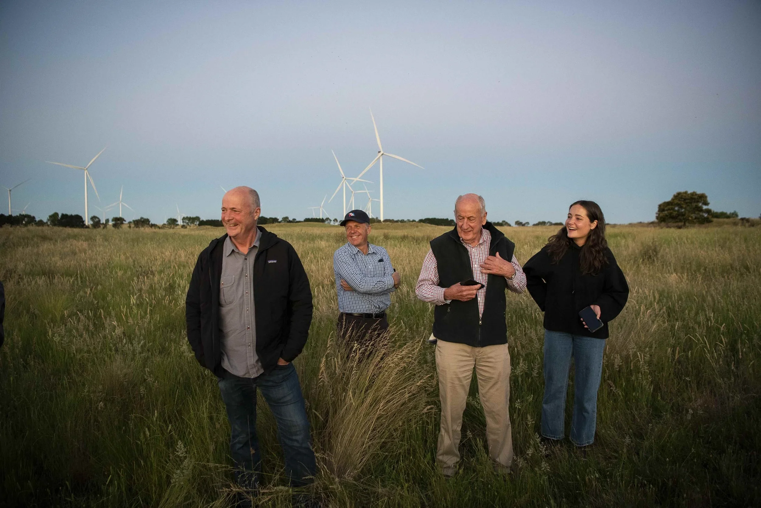 Four people in casual clothing standing and smiling in a grassy field with wind turbines in the background.