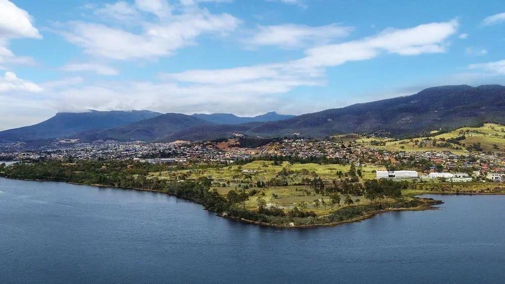 A panoramic view of a lake in the foreground, with green hills and mountains in the background under a partly cloudy sky.