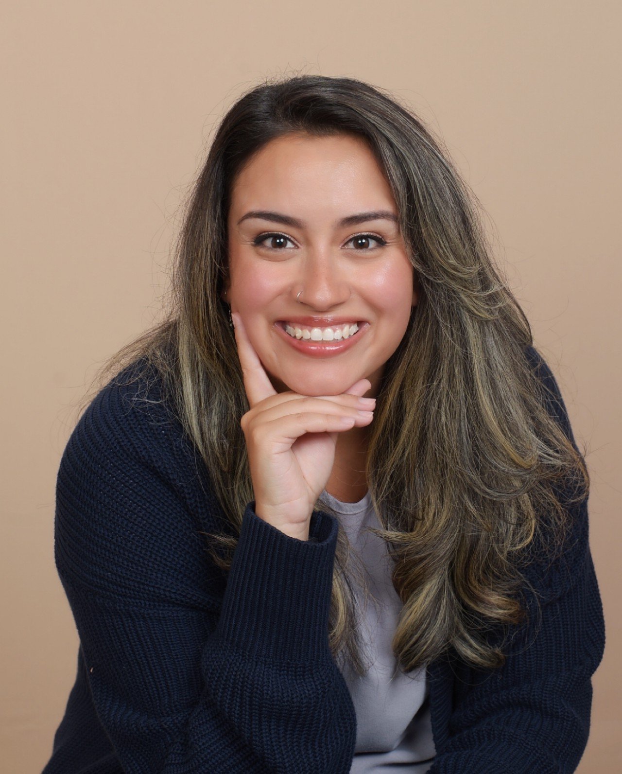A woman with long wavy hair smiling, resting her chin on her hand, wearing a black sweater and white shirt, against a beige background.