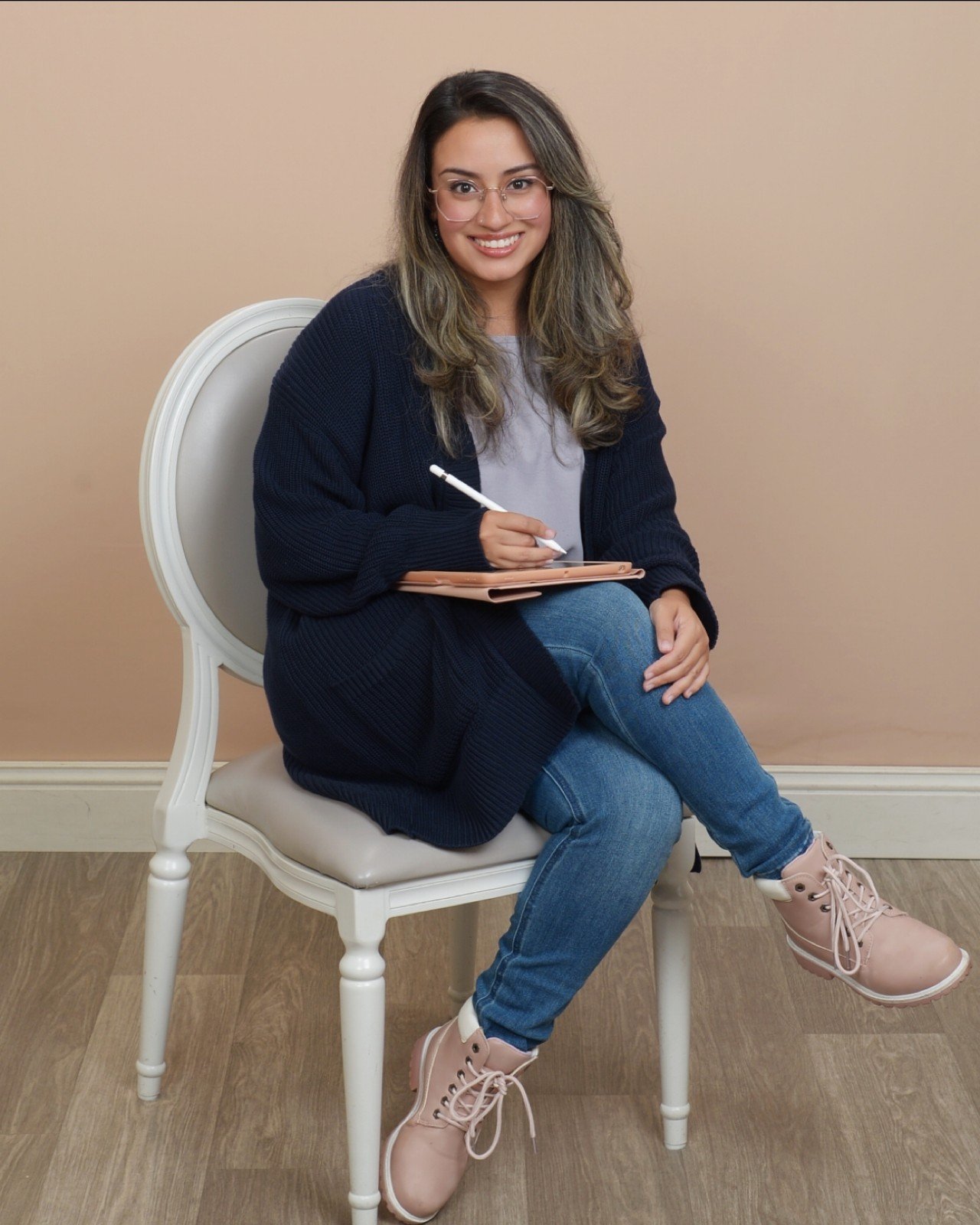A young woman with glasses and wavy hair, smiling, sitting on a white chair, holding a notepad and pen, wearing a navy cardigan, grey shirt, blue jeans, and pink boots, seated against a beige wall.