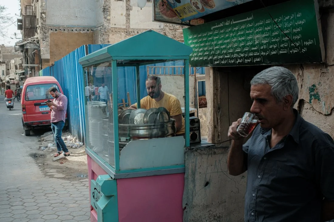 Street scene with a man in a pink checkered shirt walking on the sidewalk and looking at his phone, a red van parked near the curb, a food or snack cart with a man behind it, and a man with gray hair drinking from a glass.
