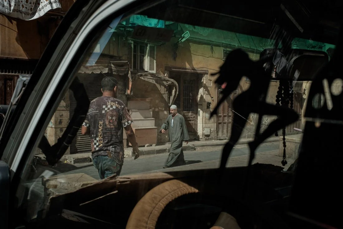 Two men walking on a street seen from inside a vehicle, with old buildings in the background.