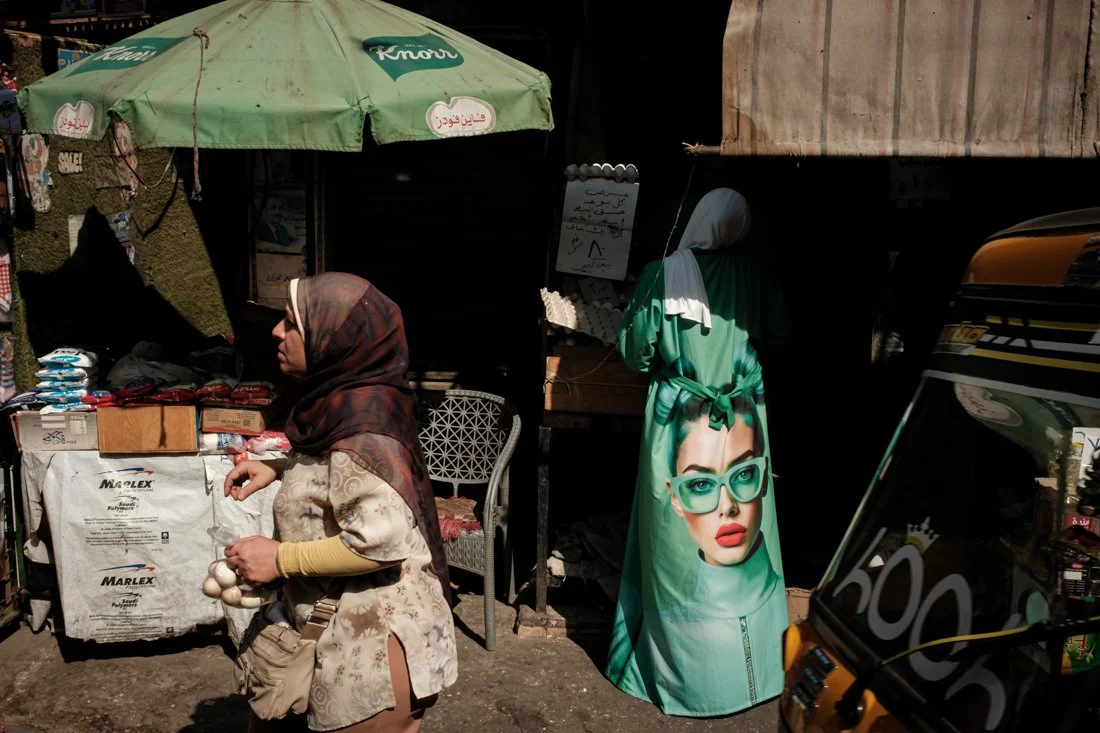 A woman wearing a headscarf is shopping at an outdoor market stall with a green umbrella. There are eggs in her hand and various items on the stall, with a chair nearby. A large blue-green dress with an image of a woman wearing glasses hangs on the s