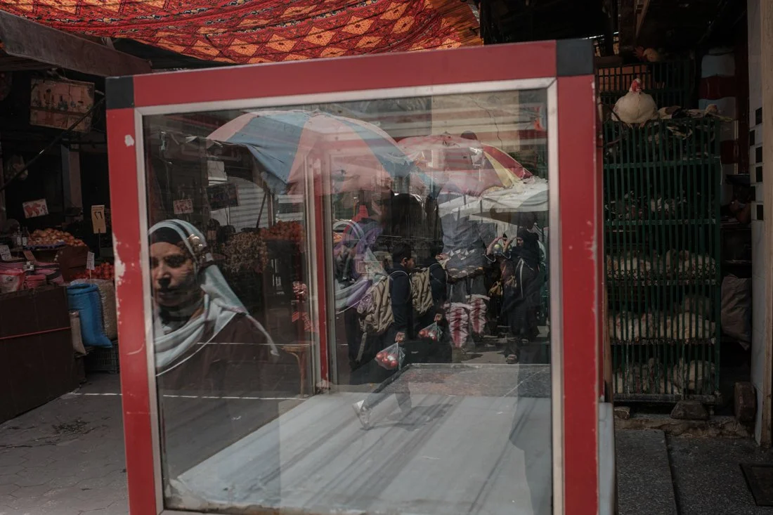 Reflection of a woman wearing a headscarf and sunglasses in a glass display case at a busy market with people shopping and vendors selling goods under umbrellas.