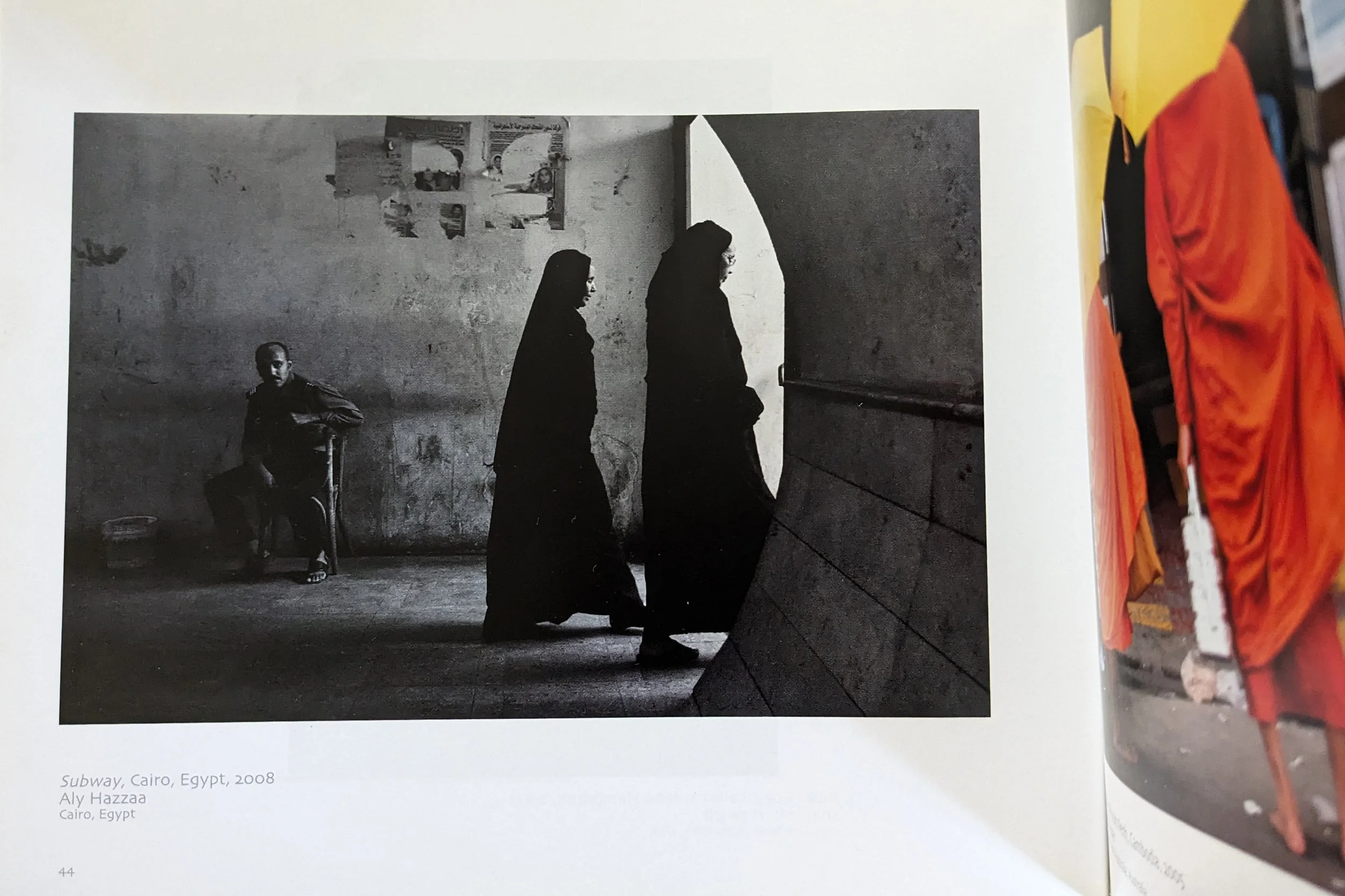 Black and white street photograph showing two women walking past a man sitting on a chair against a wall with posters in Egypt.