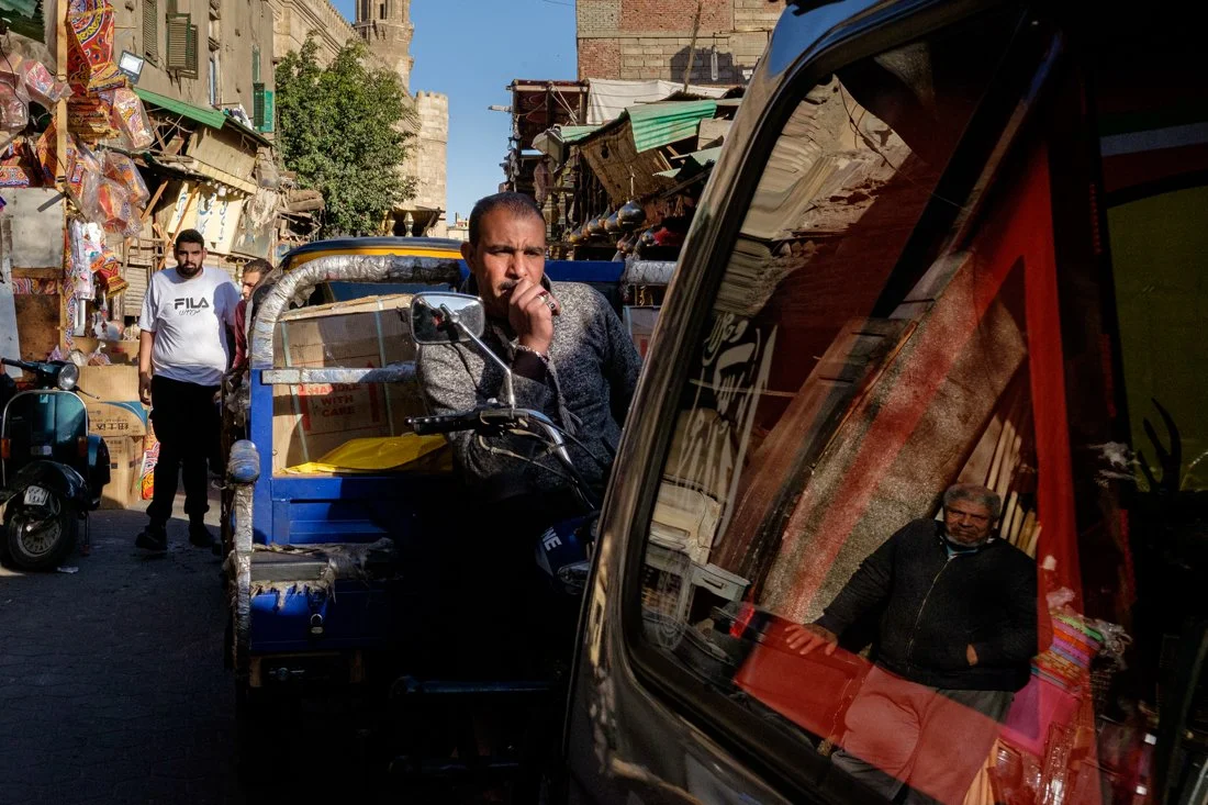 A street scene in an urban marketplace with three men, one sitting on a small vehicle and two standing nearby, surrounded by shops and stalls.