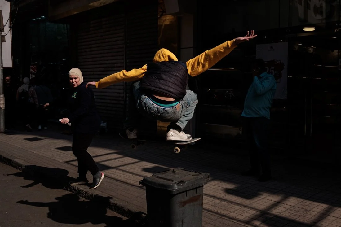 A skateboarder mid-air performing a trick on a city sidewalk with onlookers nearby, including a woman in a headscarf and a person talking on a phone, in front of closed storefronts.