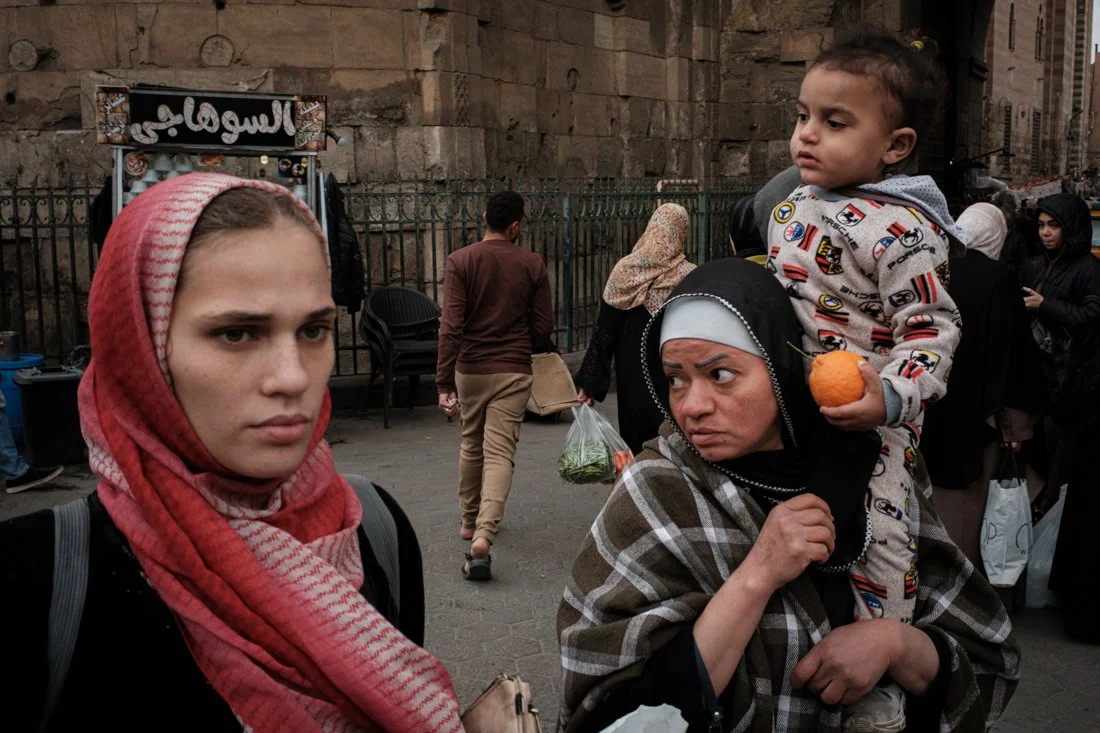 A young girl and a woman with a boy on her shoulders are in a busy outdoor market in front of a stone building and iron fence. The girl has a red patterned scarf, and the woman wears a black and white headscarf. The boy holds an orange ball and wears