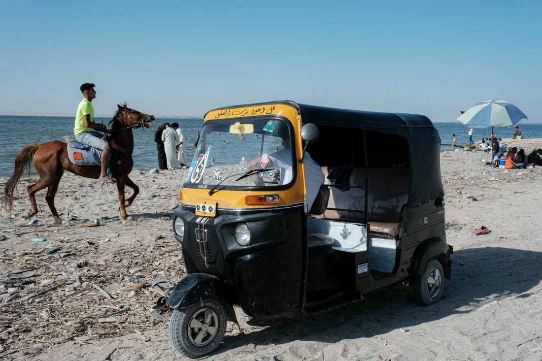 A black and yellow auto rickshaw parked on a beach with a view of the sea, people walking along the shoreline, a person riding a horse, and a group sitting under an umbrella.