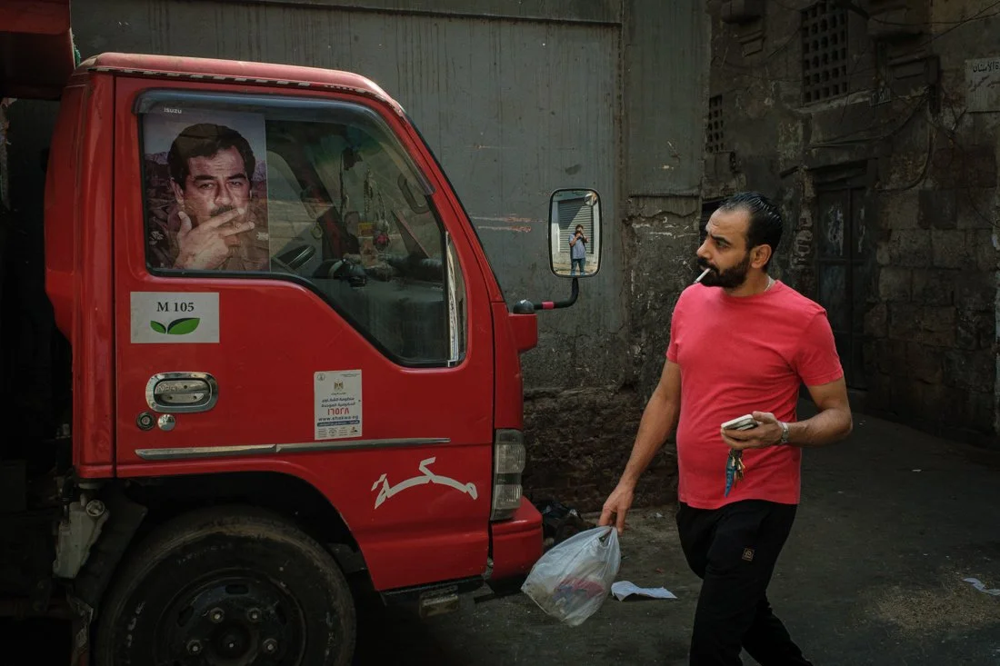 A man in a red t-shirt walks past a small red truck with an advertisement featuring a man's face, holding a cigarette. The man has a cigarette in his mouth and is holding a phone in one hand and a plastic bag in the other. The reflection of the man t